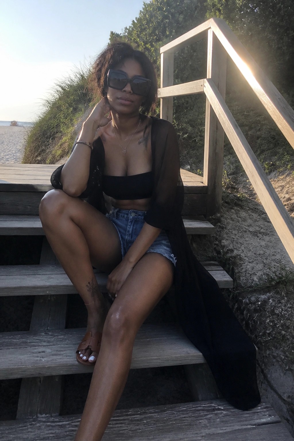Woman in sheer black top and denim shorts sits on sunny beach stairs wearing sunglasses