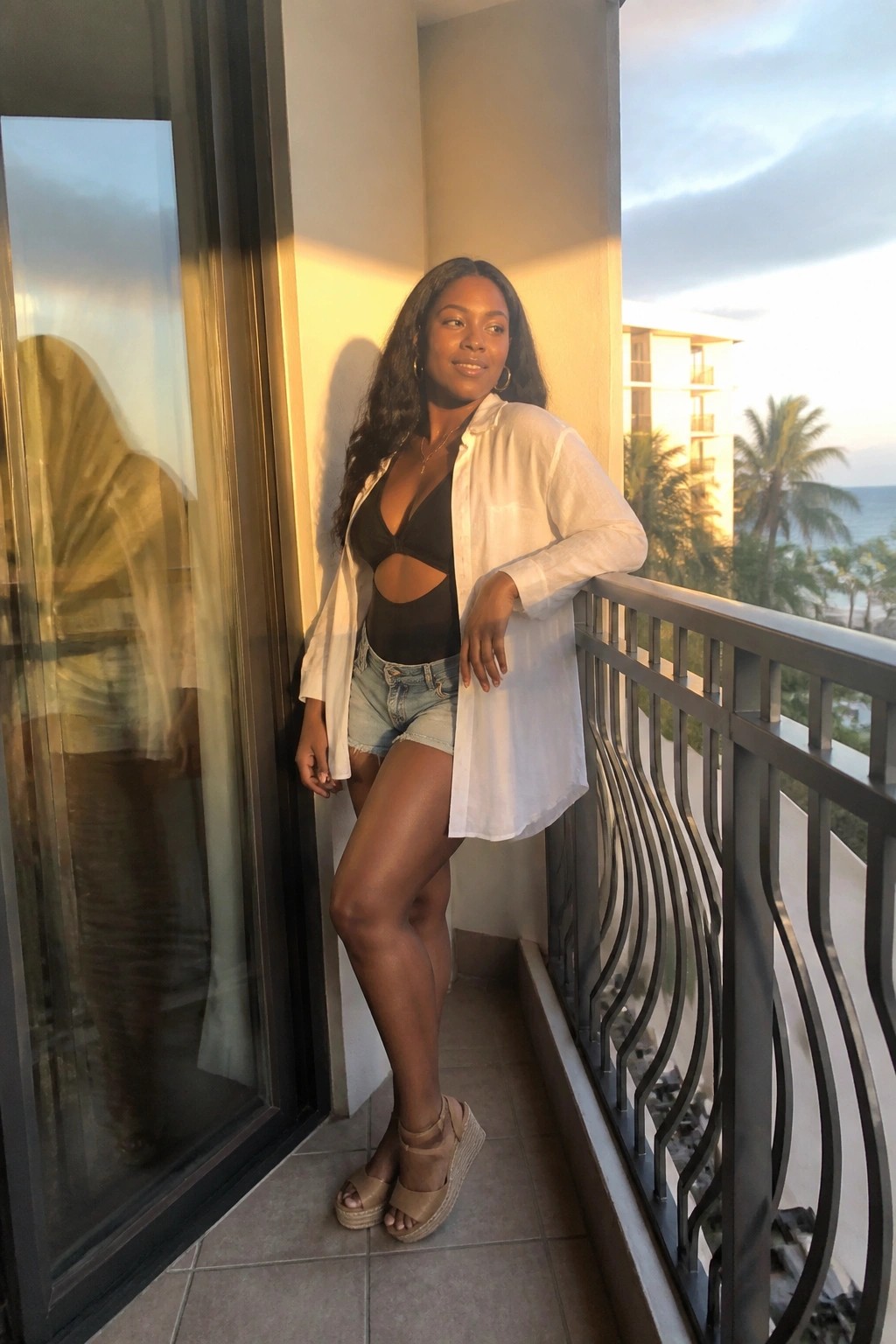 Woman in white shirt over black cutout swimsuit on ocean-view balcony at sunset