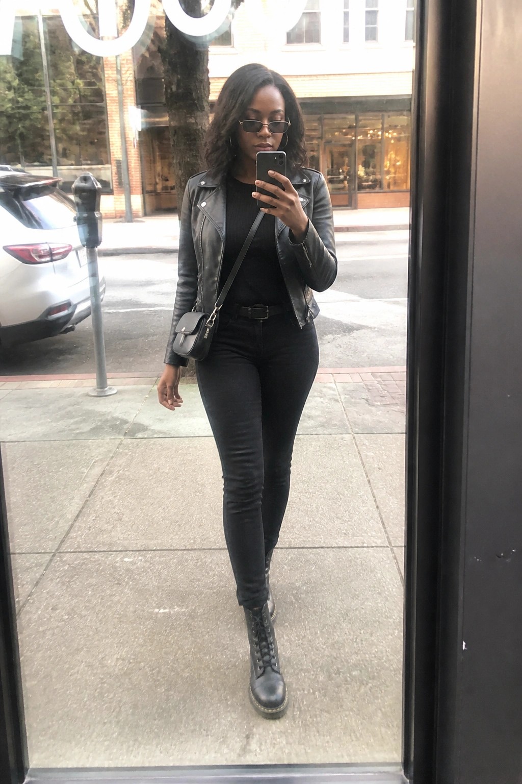 Woman in black leather jacket taking mirror selfie on city sidewalk with phone