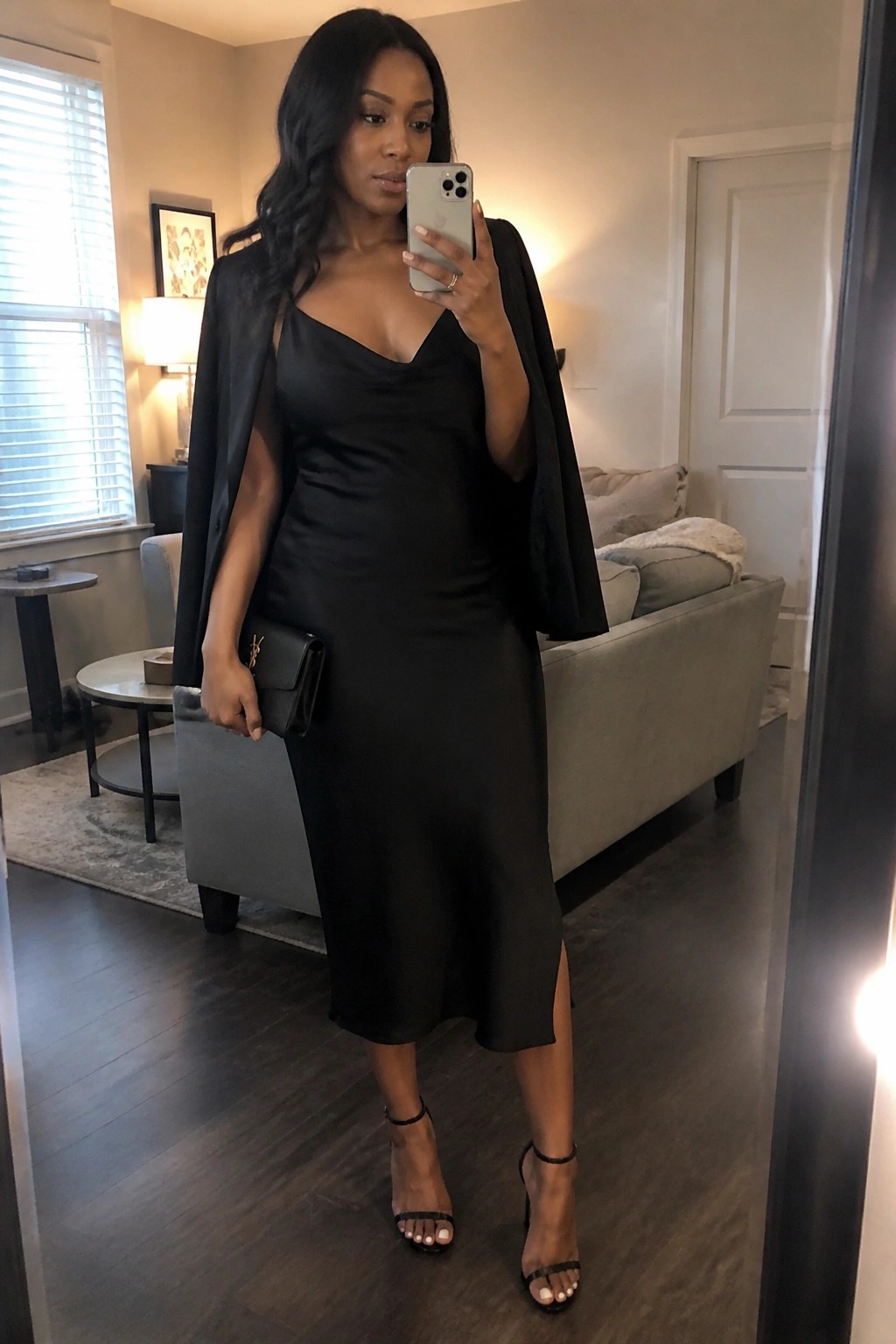 Woman wearing a black satin midi slip dress with a black blazer draped over her shoulders, holding a black clutch and smartphone, paired with black strappy heels in a monochrome outfit.