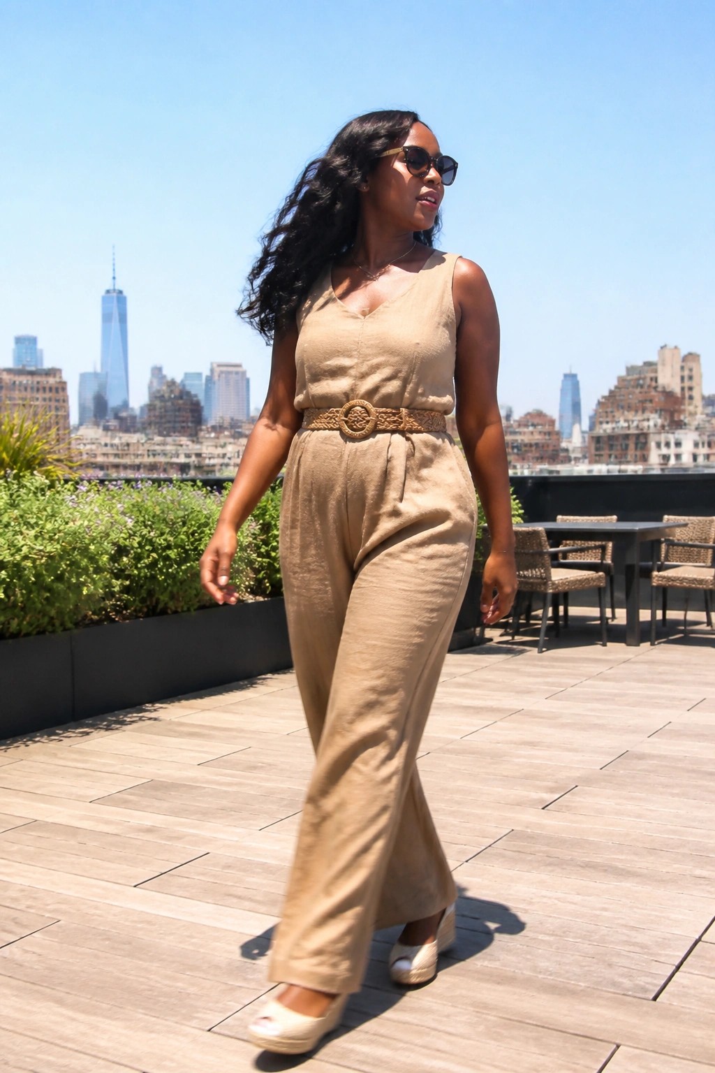 Woman in beige jumpsuit and sunglasses walks on sunny rooftop with city skyline.
