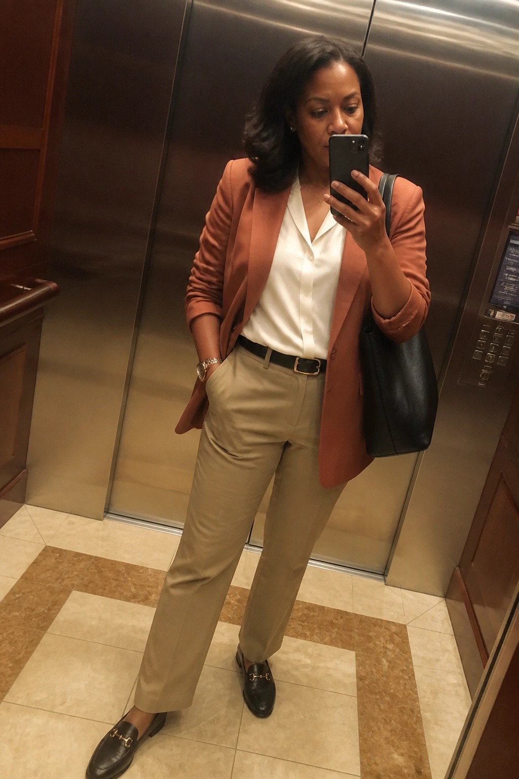 Woman in rust blazer, white blouse, and beige pants taking elevator mirror selfie