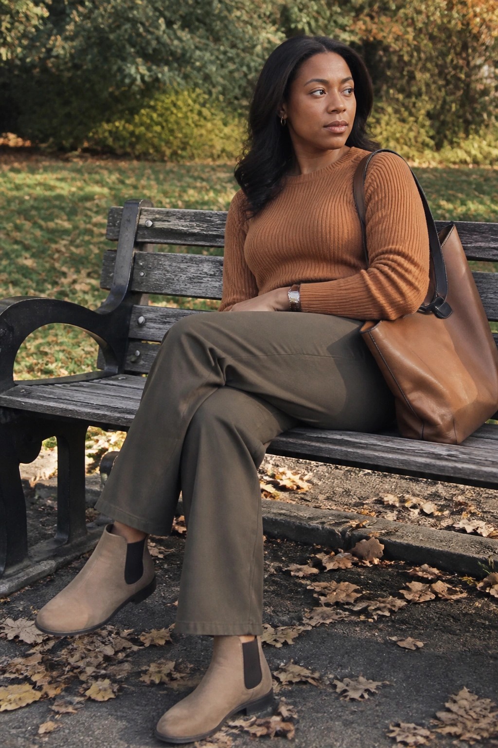 Black woman seated on a park bench wearing a brown ribbed knit sweater, olive wide-leg pants, taupe suede Chelsea boots, and carrying a brown leather tote bag in an earth tone outfit.
