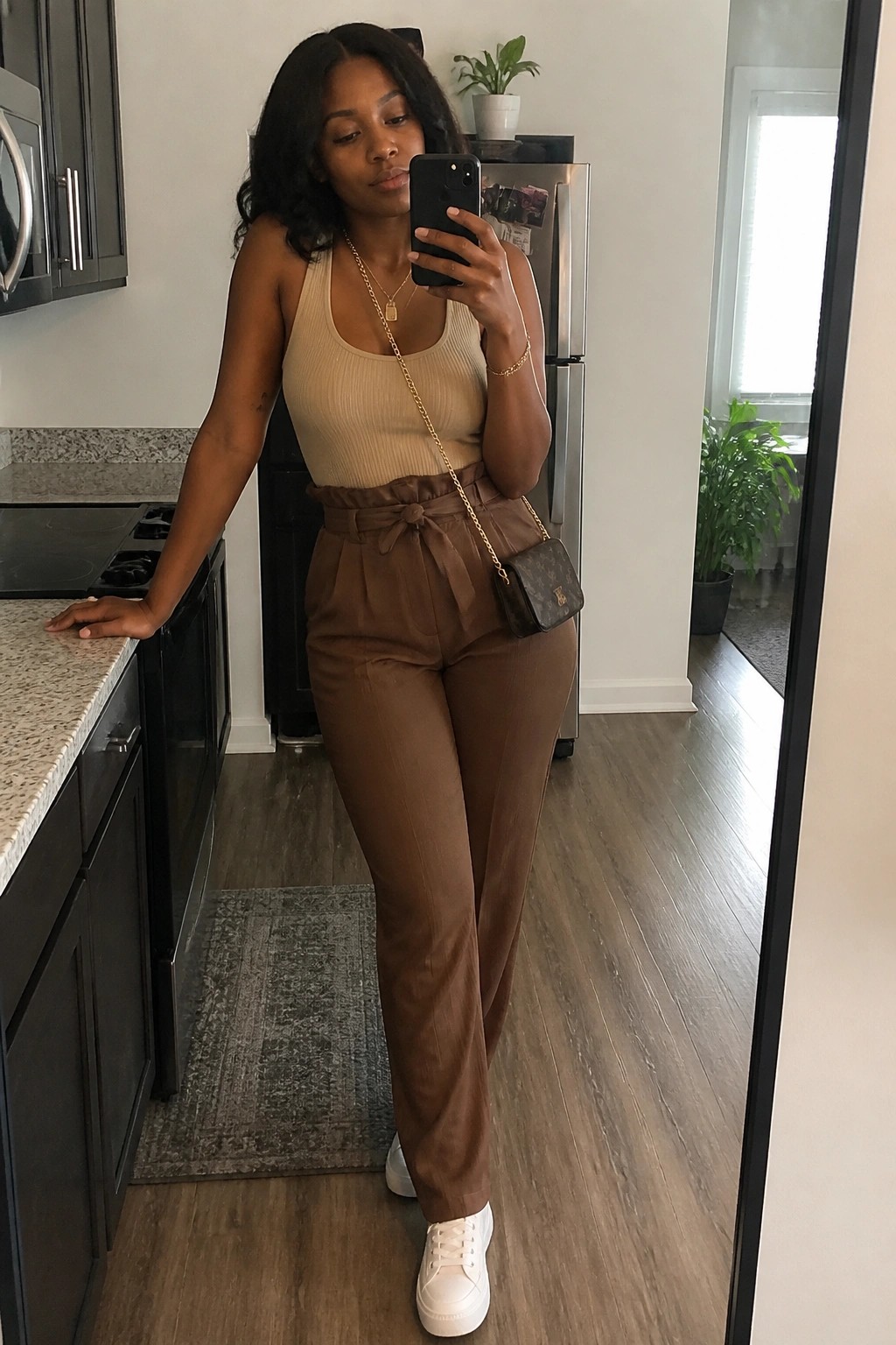 Woman in beige tank top and brown belted pants taking kitchen mirror selfie