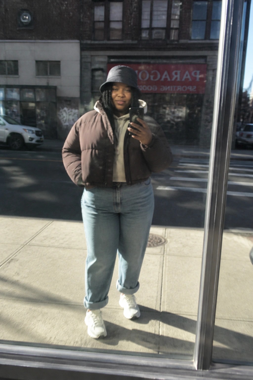 Woman in brown puffer jacket and bucket hat taking phone selfie on sidewalk