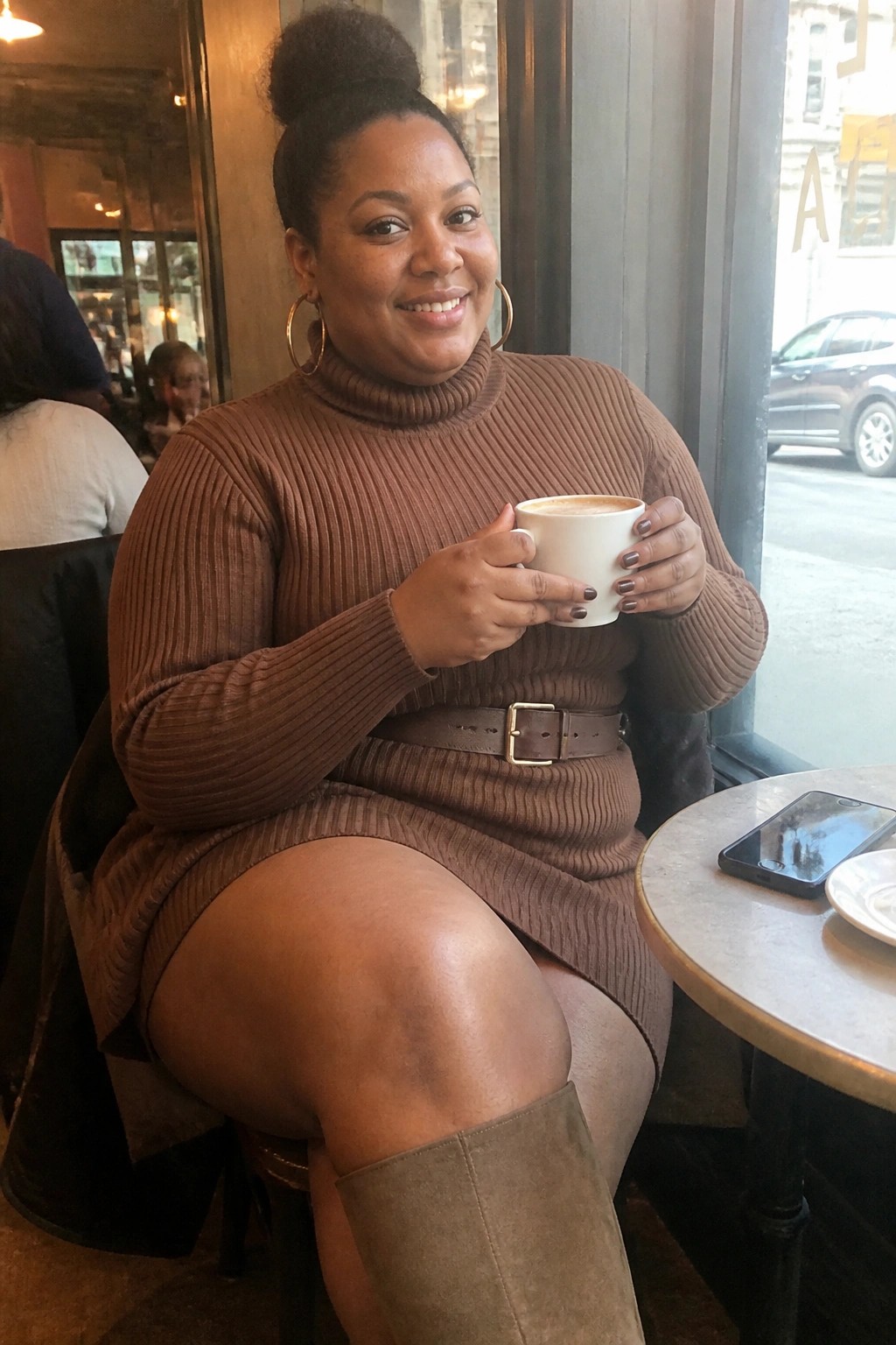 Smiling woman in brown turtleneck sweater dress holding coffee in cafe