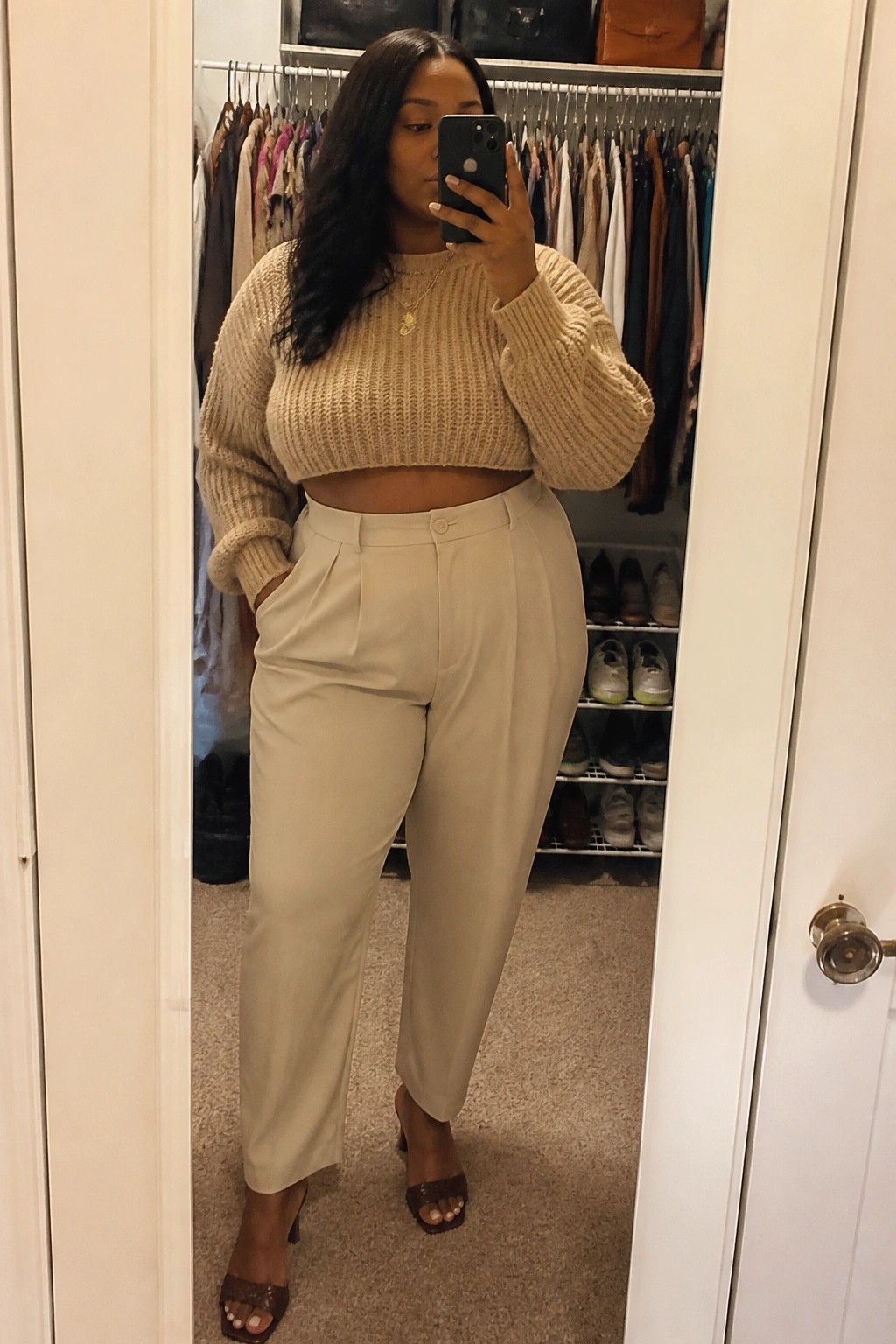 A plus size Black woman taking a mirror selfie in a beige cropped chunky knit sweater, matching pleated trousers, and brown heeled sandals, standing in a closet.
