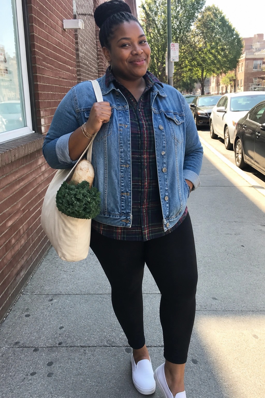 Plus size Black woman in a blue denim jacket over a plaid shirt, black leggings, and white slip on shoes while carrying a tote bag with vegetables.