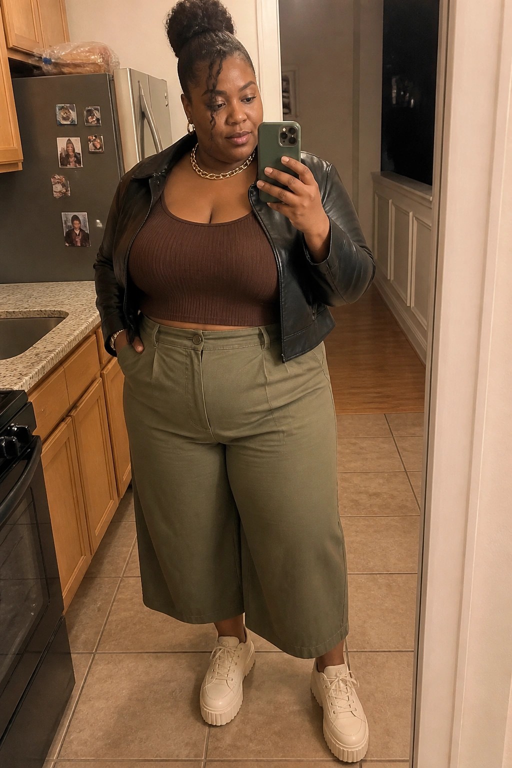 Woman in brown crop top, black jacket, and green pants taking mirror selfie in kitchen