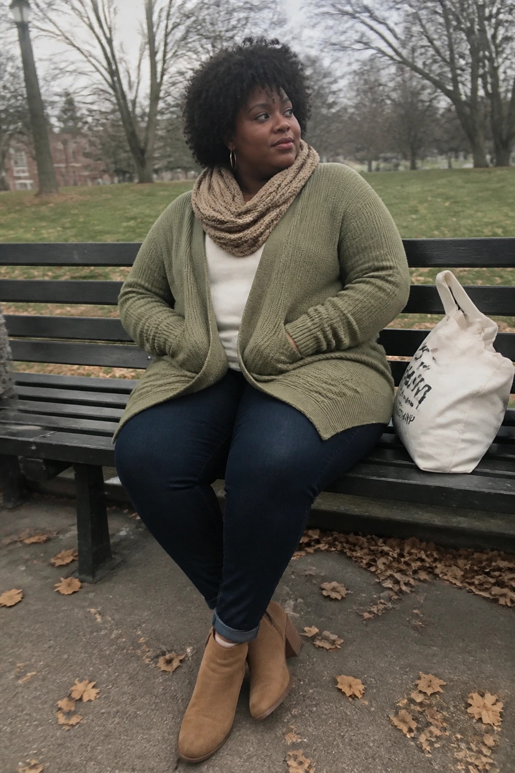 Woman in green cardigan and beige scarf on park bench beside tote bag