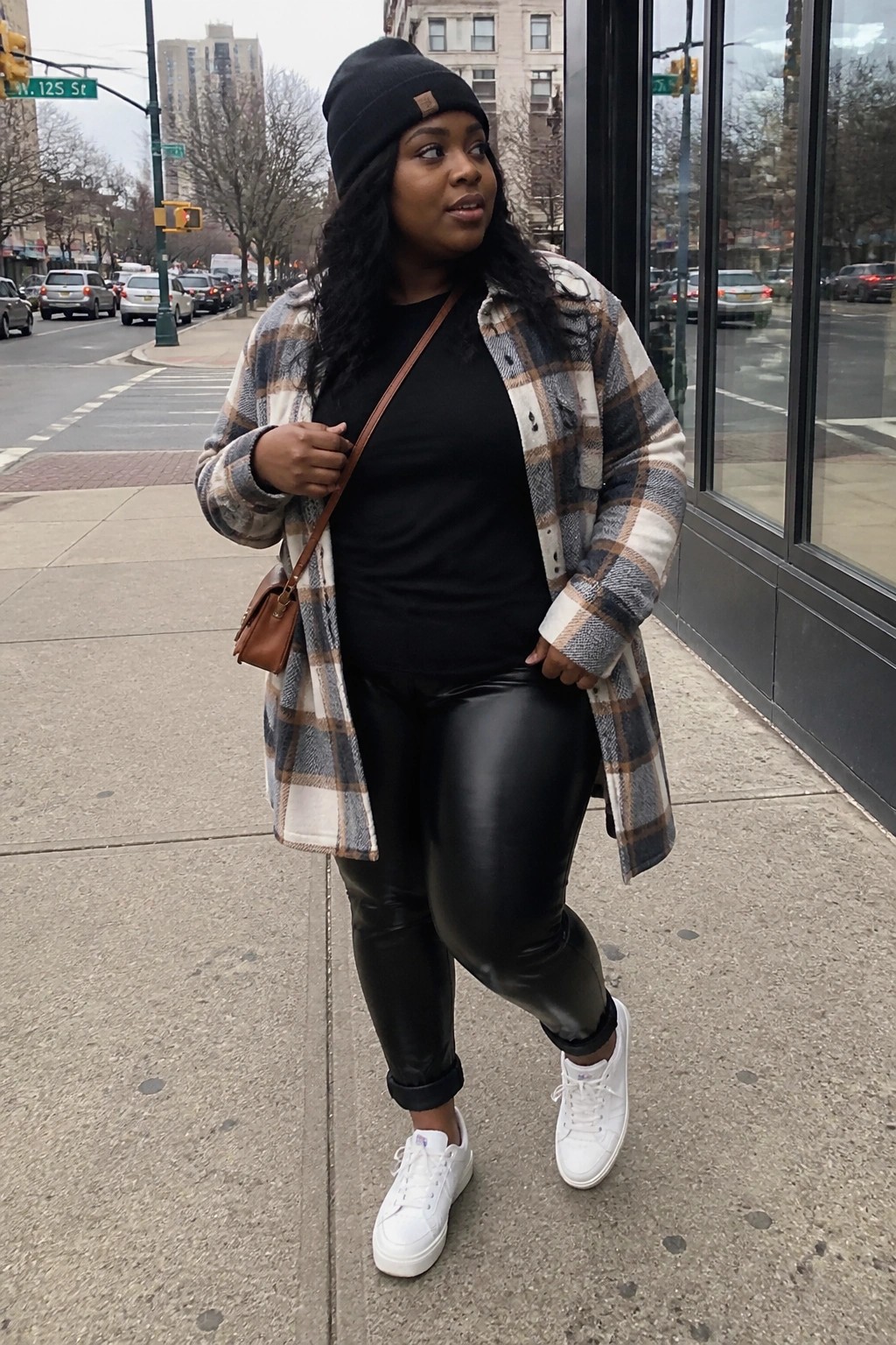 Plus size Black woman wearing a long brown and white plaid shacket open over a black tee, black faux leather leggings, white sneakers, black beanie, and brown crossbody bag on a city sidewalk.