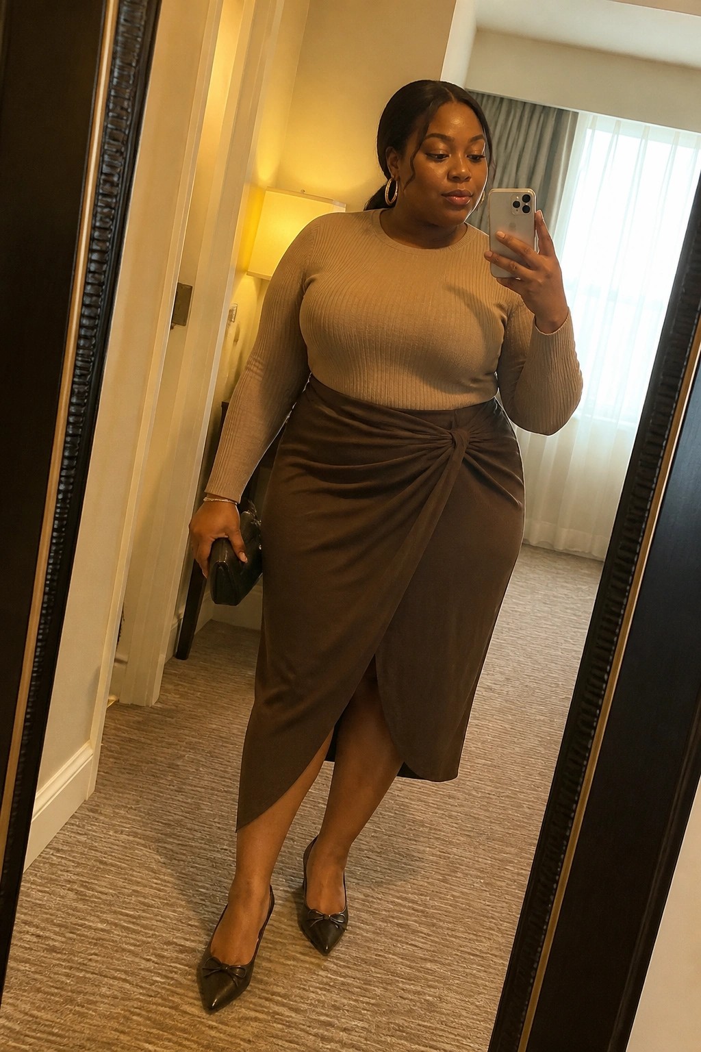 Plus size woman wearing a beige ribbed long sleeve top tucked into a brown wrap midi skirt with front knot detail, black pointed flats with bows, gold hoop earrings, and holding a black clutch while taking a mirror selfie.