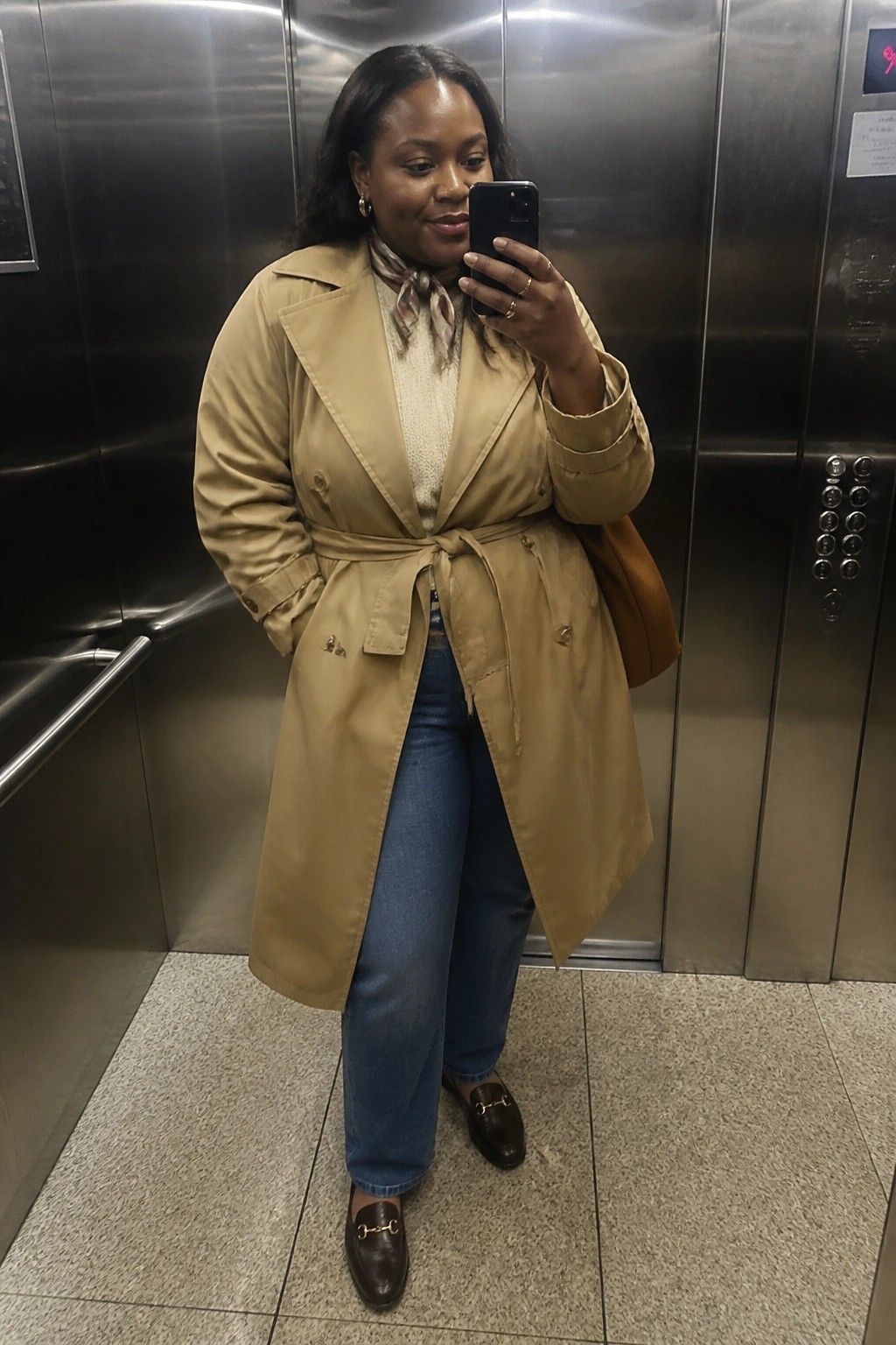 Woman in tan trench coat taking selfie in stainless steel elevator