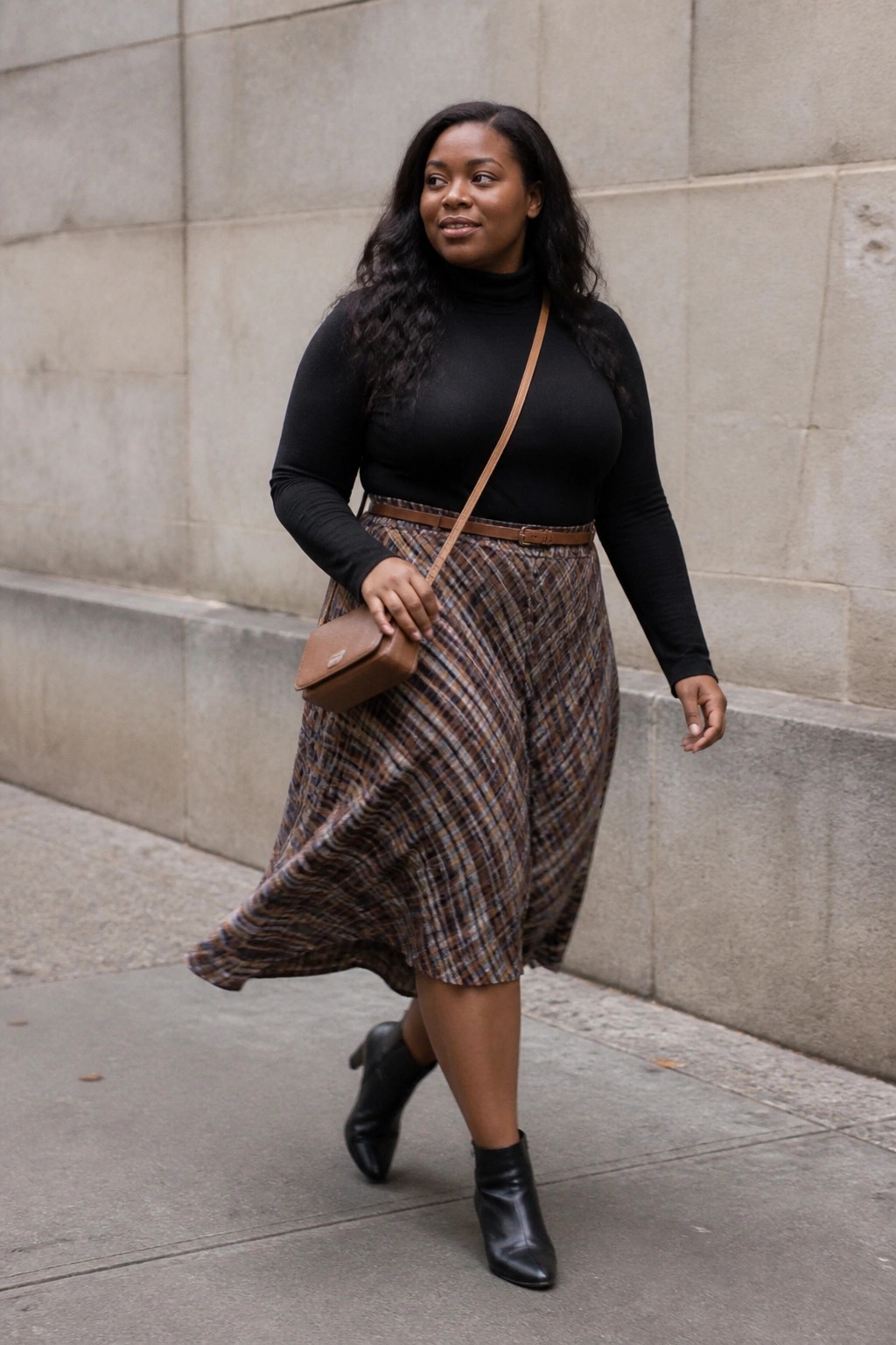 Woman in black turtleneck and flowing plaid skirt with brown crossbody bag on sidewalk