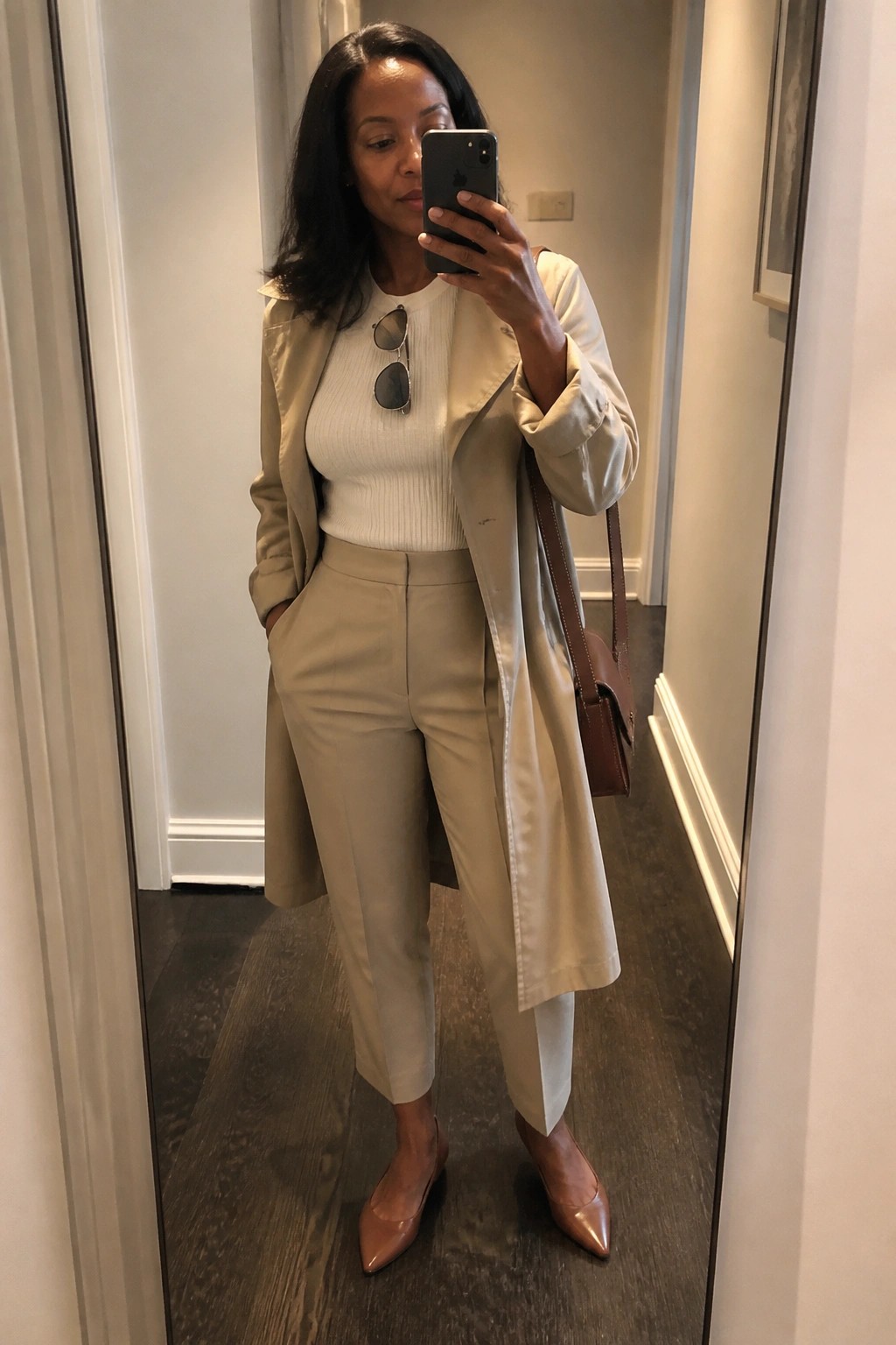Woman in beige trench coat and trousers taking mirror selfie with phone