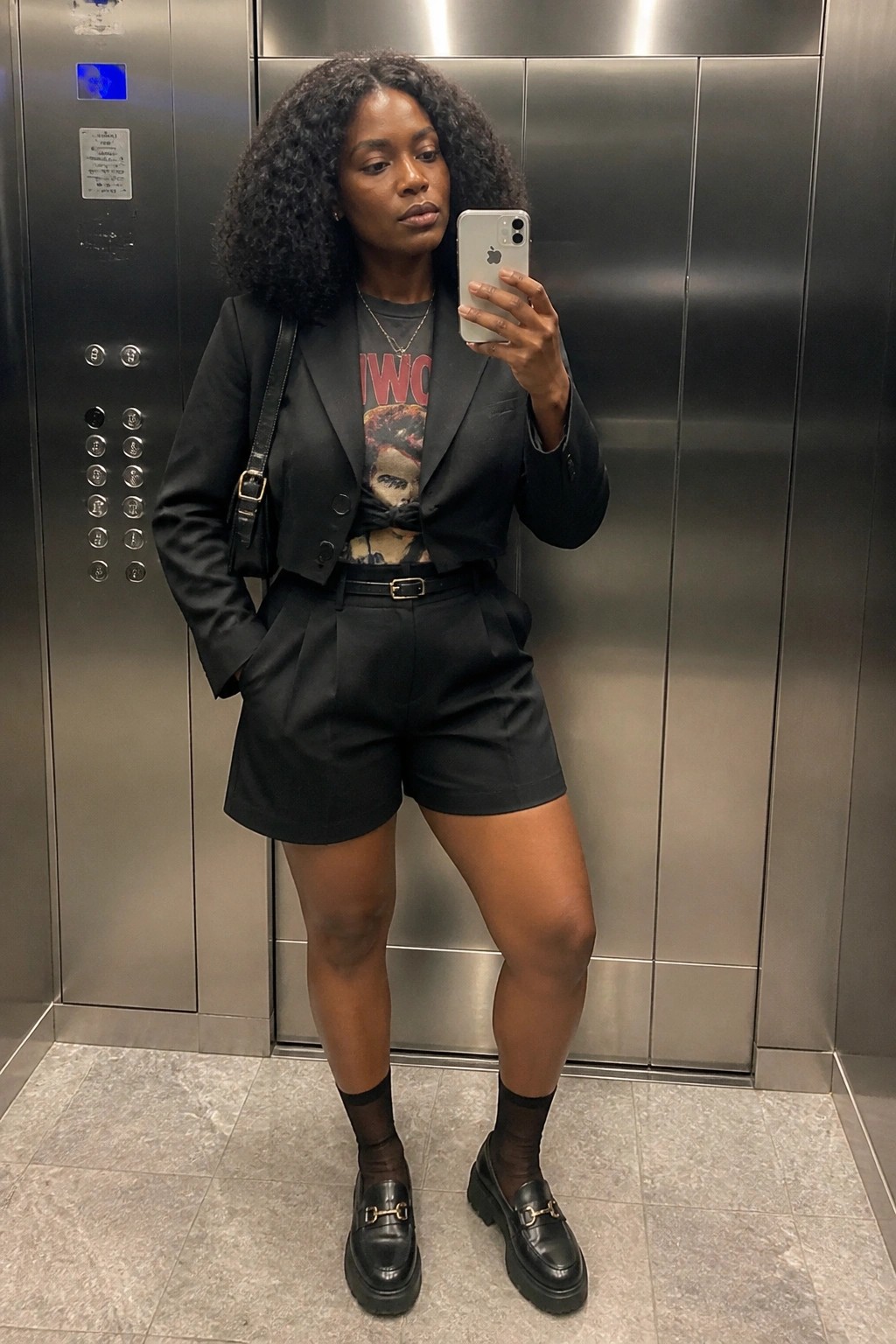 Woman with curly hair taking selfie in elevator wearing black blazer and shorts