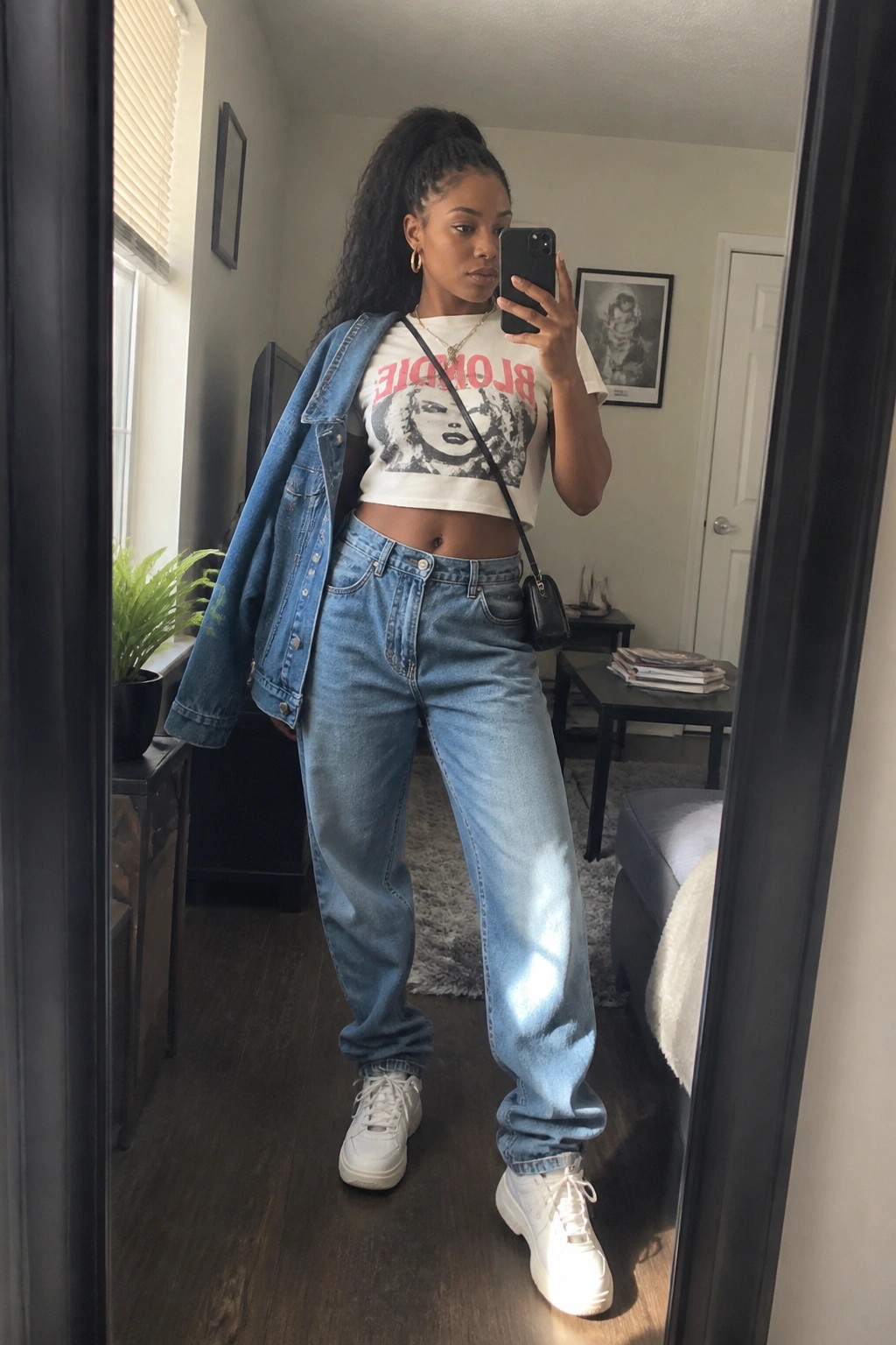 Woman taking mirror selfie in denim jacket, Blondie crop top, and light blue jeans
