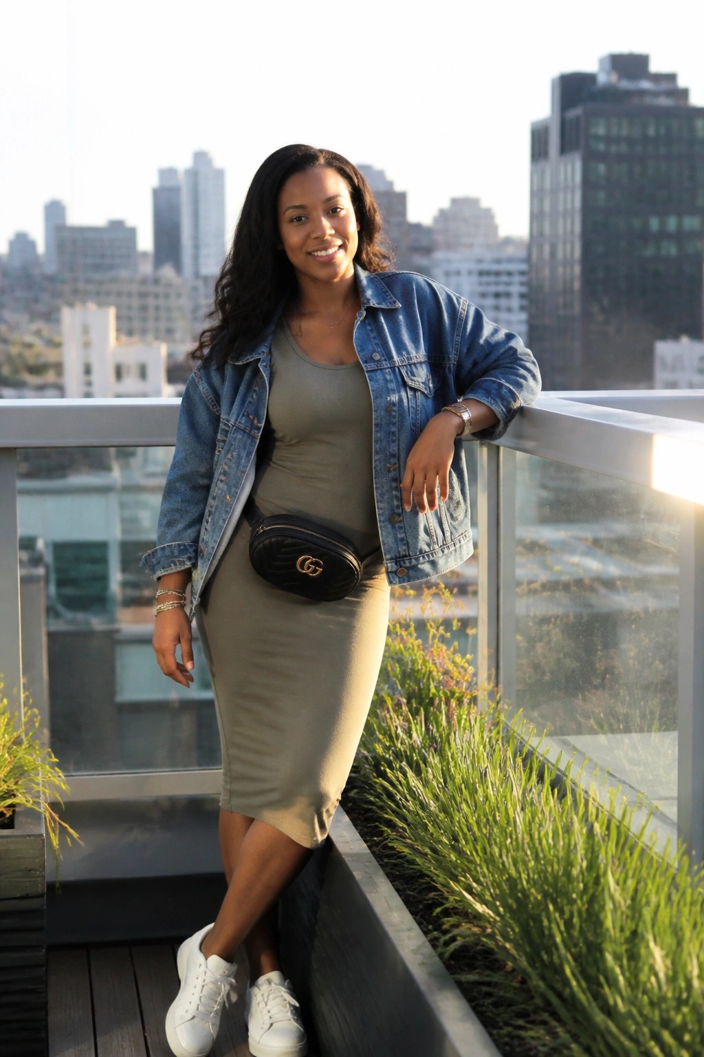 Woman in olive dress and denim jacket smiling on city rooftop balcony.