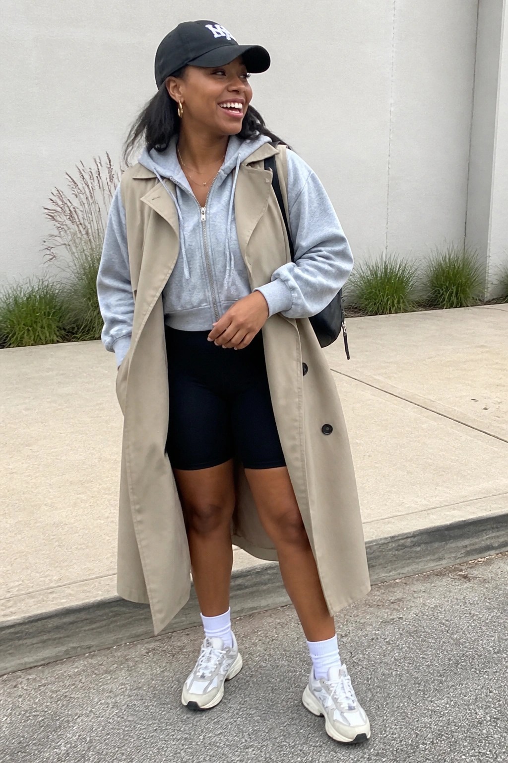 Smiling woman in black NY cap, beige trench vest, gray hoodie, and biker shorts