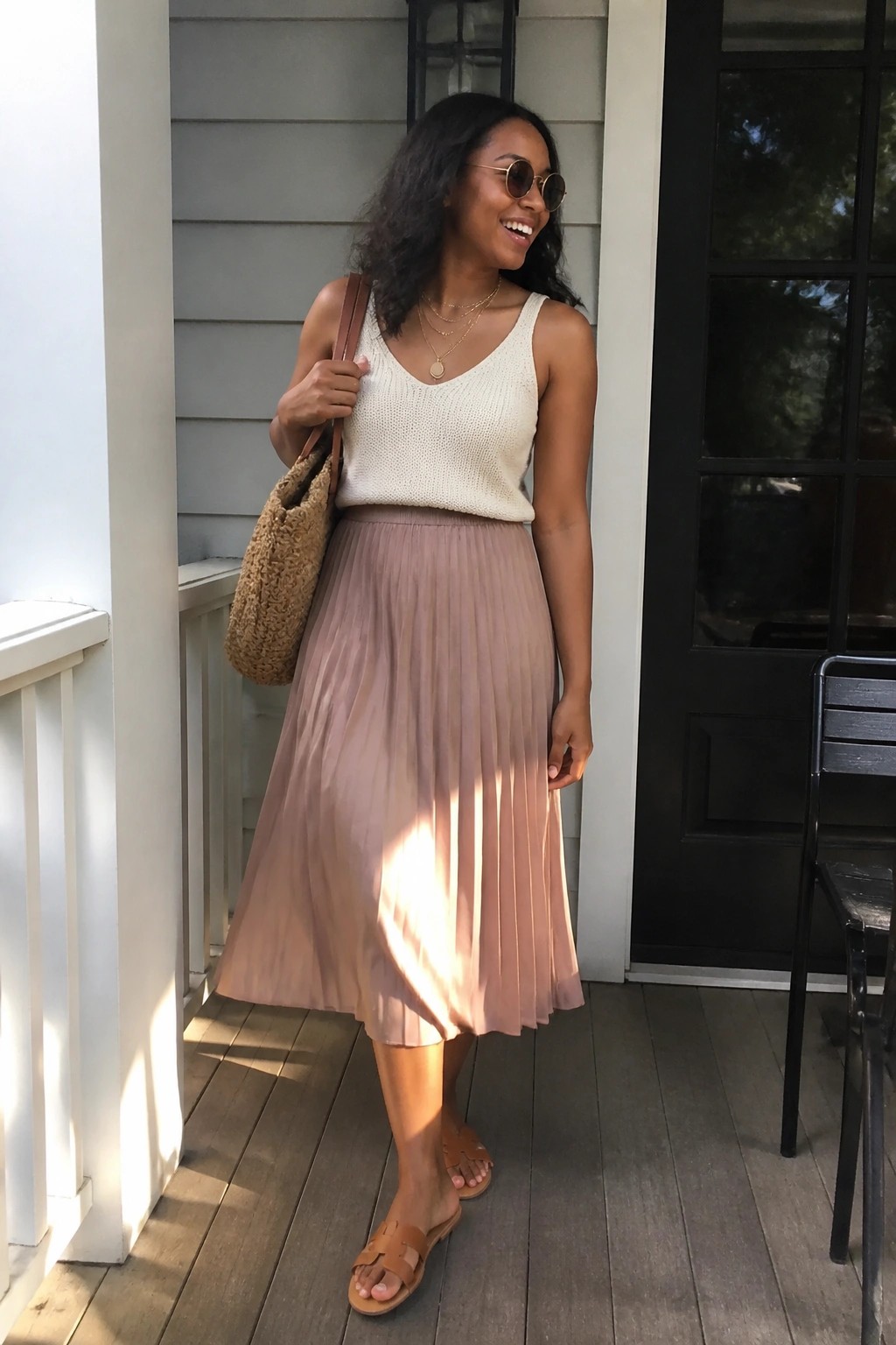 Woman in cream knit tank top, dusty rose pleated midi skirt, brown sandals, layered gold necklaces, round sunglasses, and fuzzy brown tote bag standing on a porch.