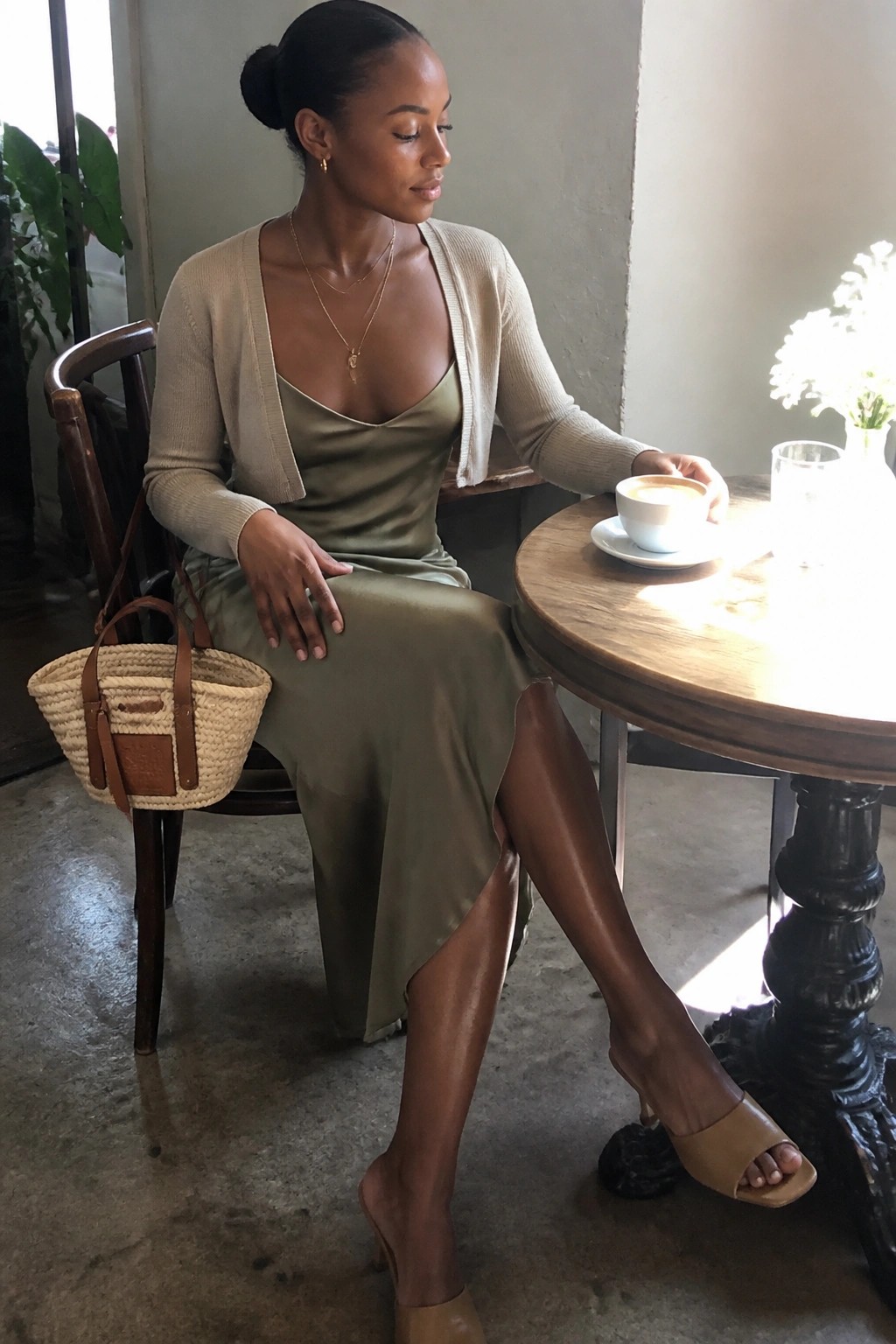 Woman in green satin dress and beige cardigan at cafe table with coffee and woven bag