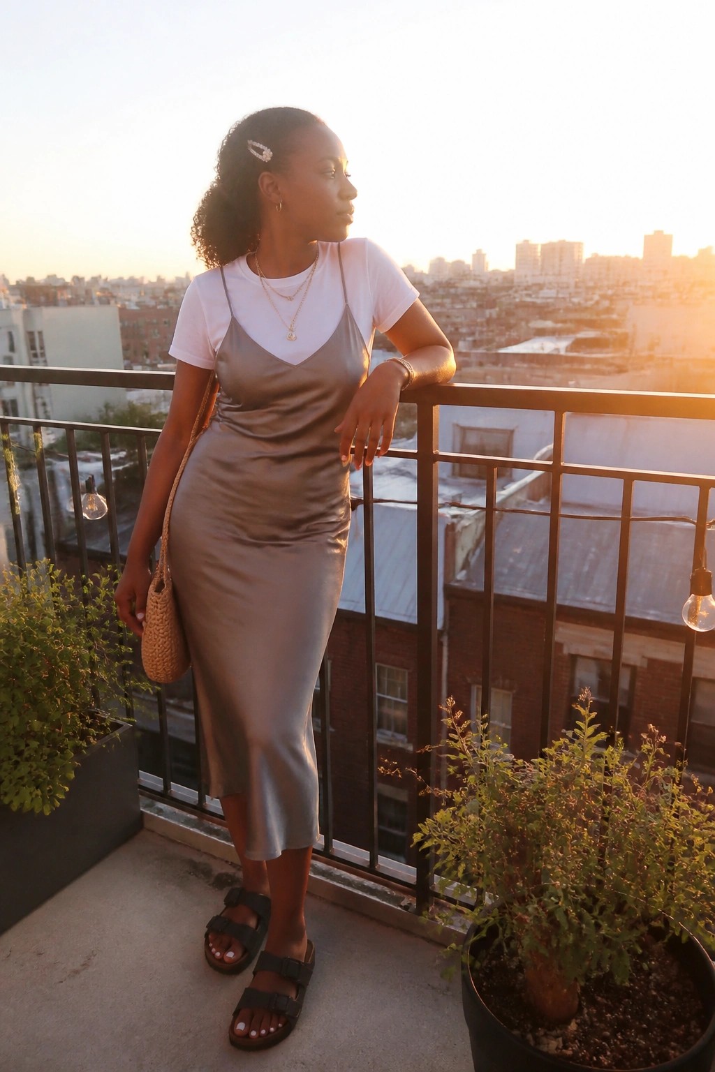 Woman in gray satin slip dress over white tee on city balcony at sunset