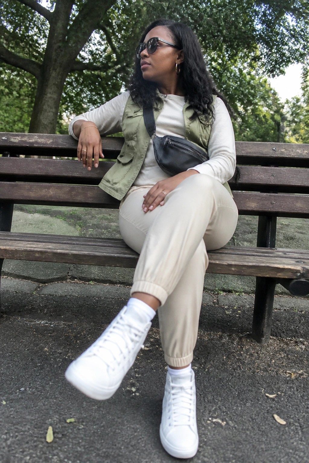 Woman in green vest, beige pants, and white sneakers seated on park bench