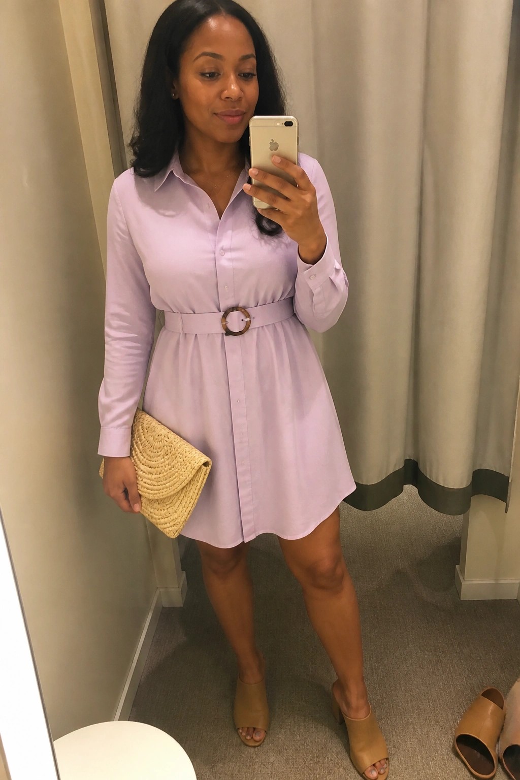 Woman wearing a lavender button-up shirt dress belted at the waist with a circular buckle, holding a woven straw clutch and wearing tan heeled mules while taking a mirror selfie.