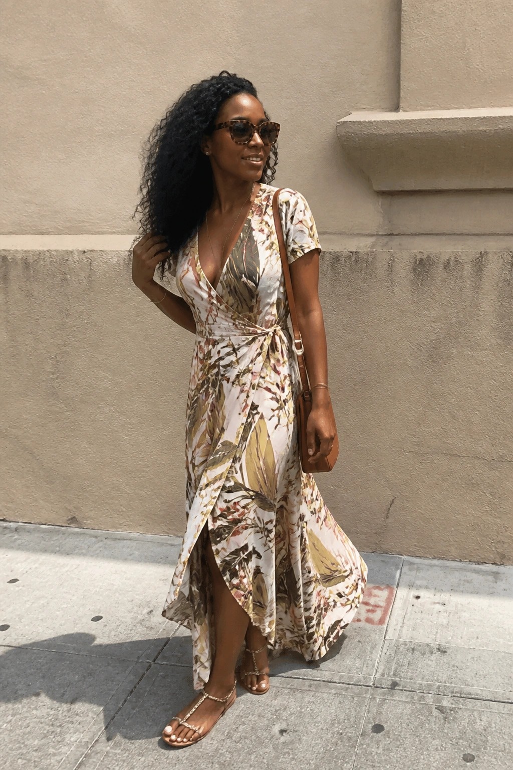 Woman in floral wrap maxi dress with sunglasses and brown bag on sidewalk.