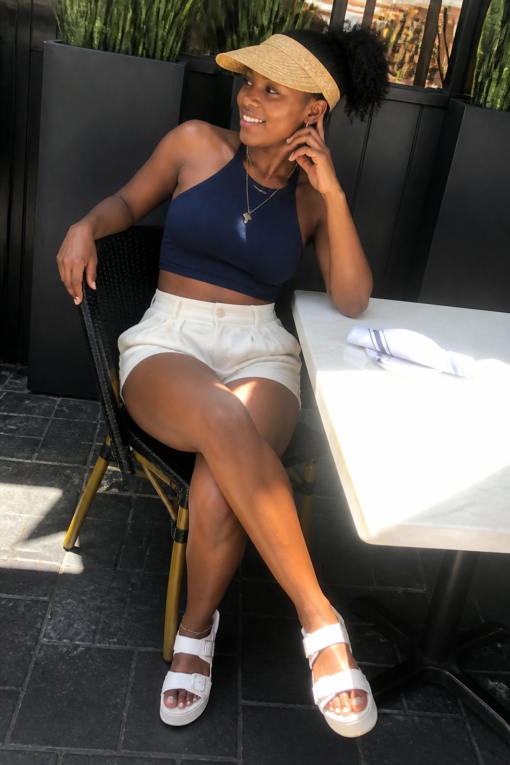Woman in navy crop top and white shorts with visor at sunny outdoor table