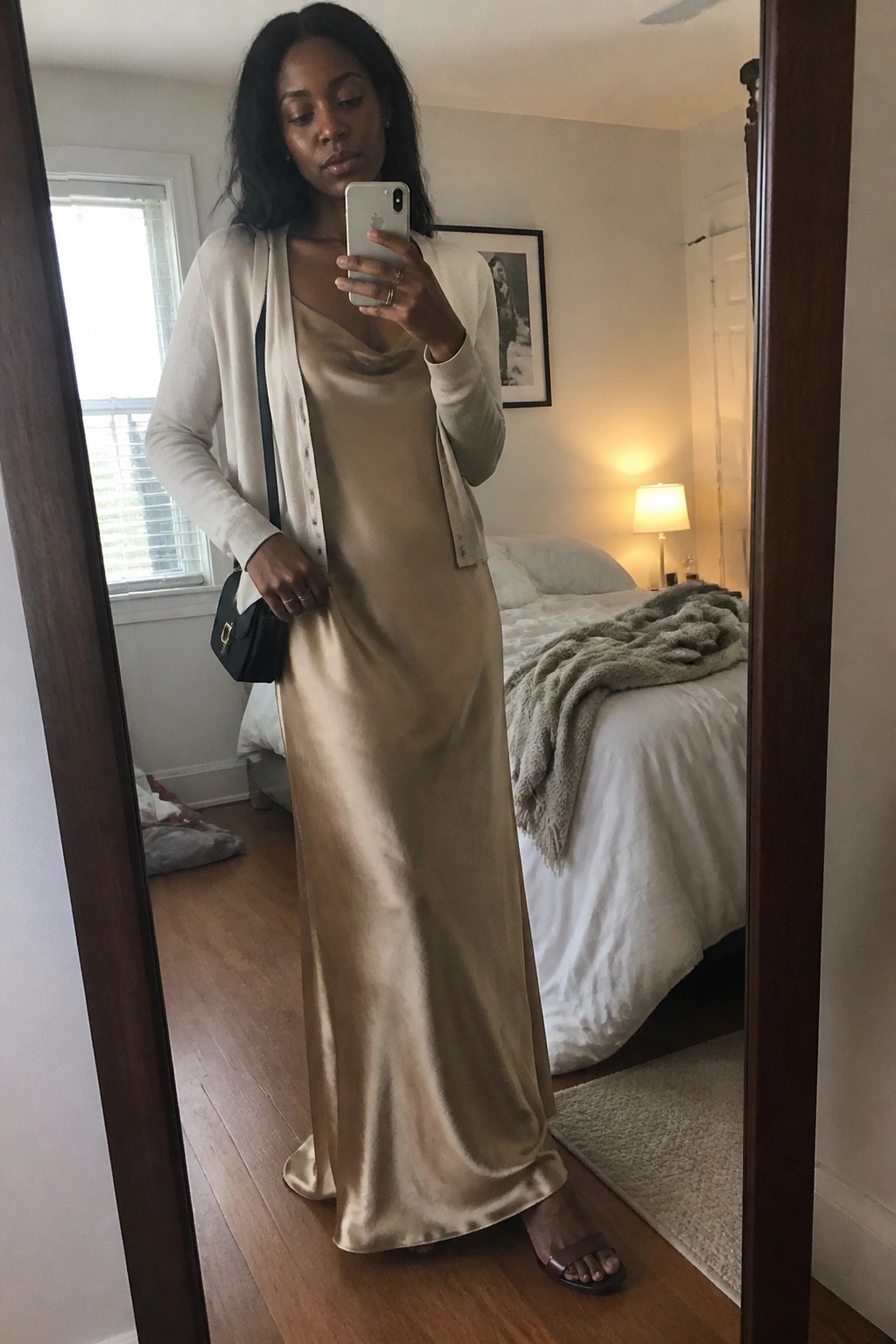 Woman in beige satin dress and white cardigan taking mirror selfie in bedroom