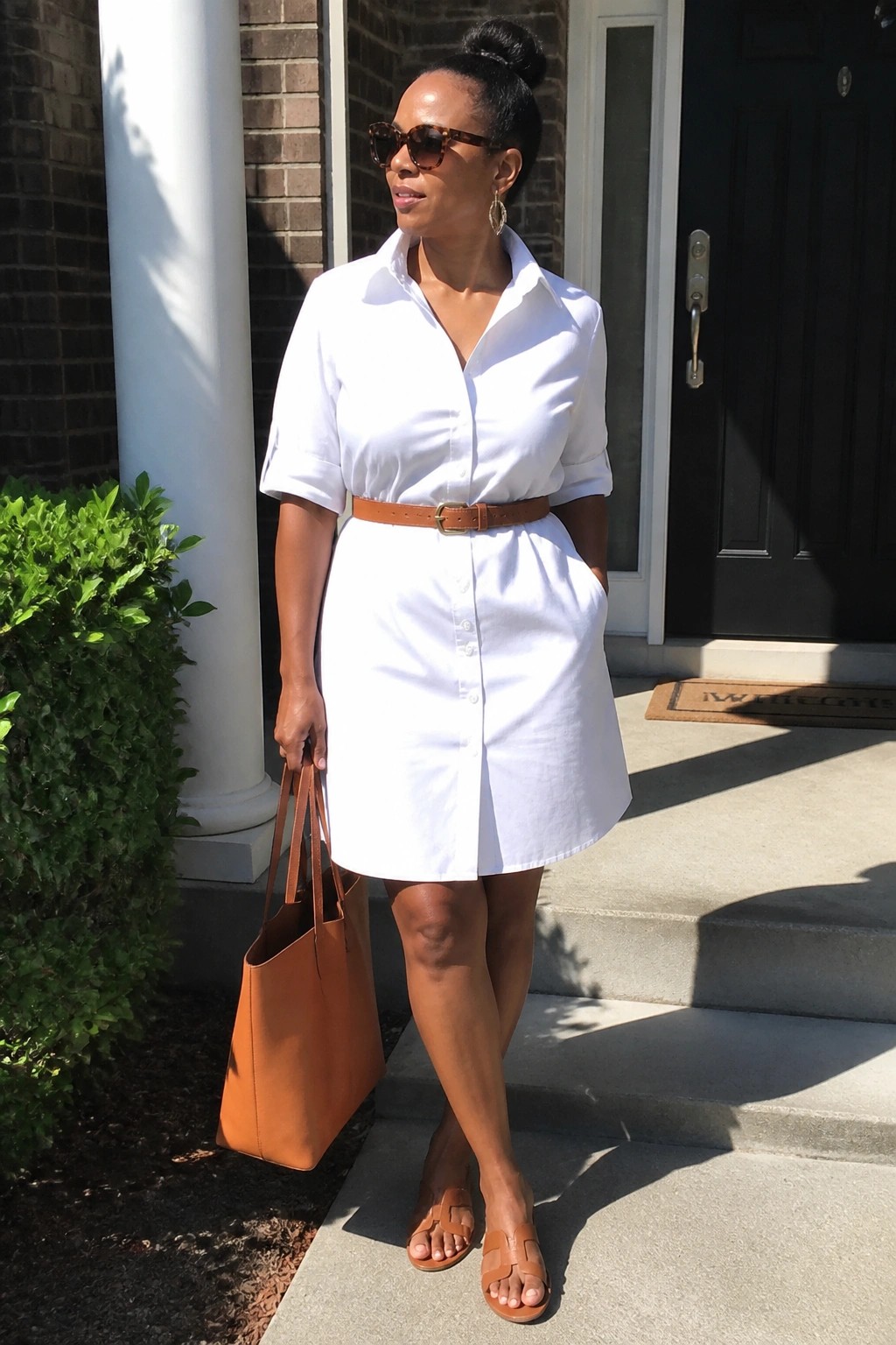 Woman in white shirt dress with brown belt holding tan leather tote outdoors