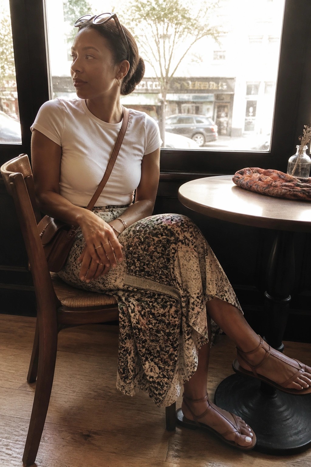 Woman in white t-shirt and floral skirt seated by cafe window with leather bag
