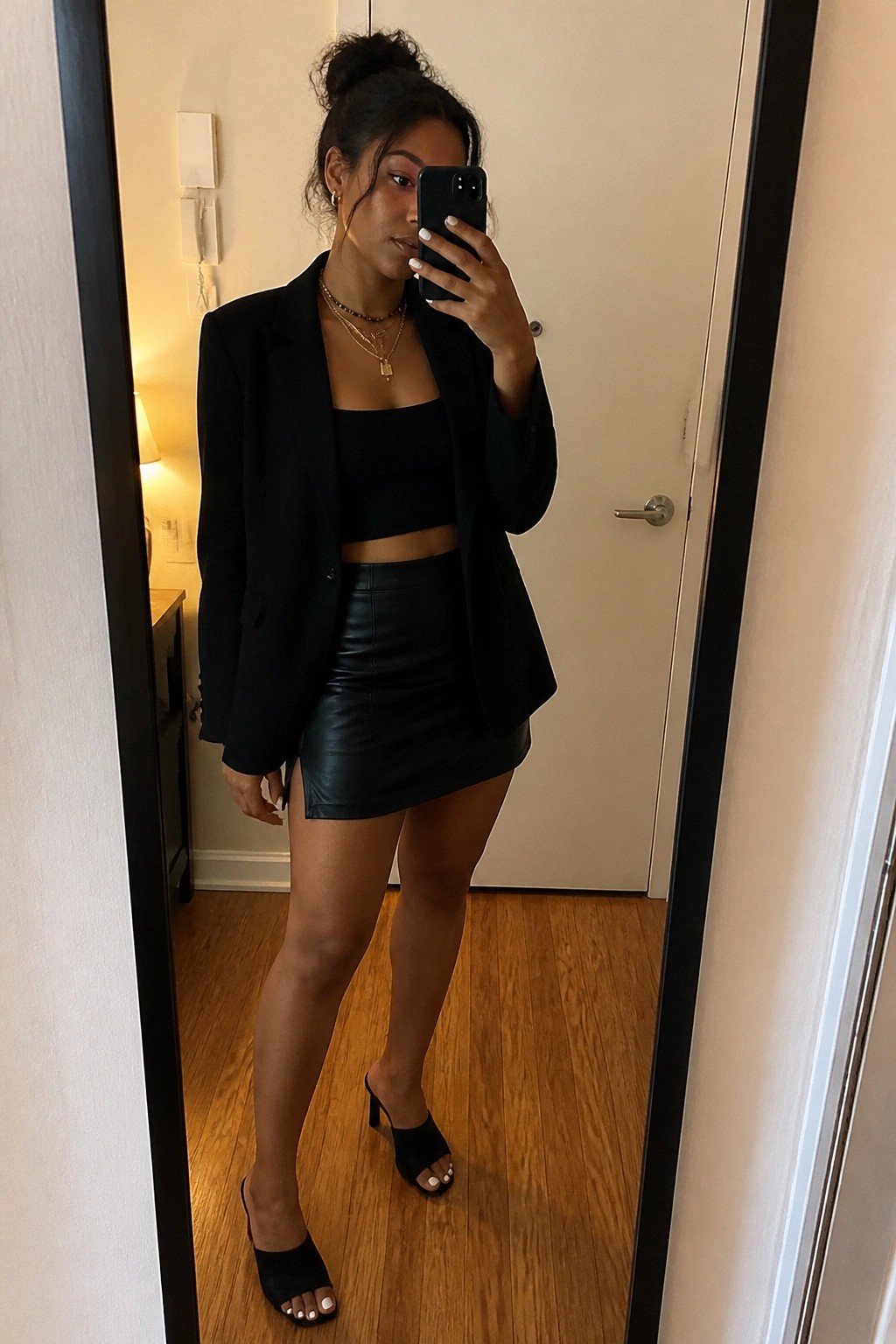 Woman in black blazer, crop top, and leather skirt taking mirror selfie with phone