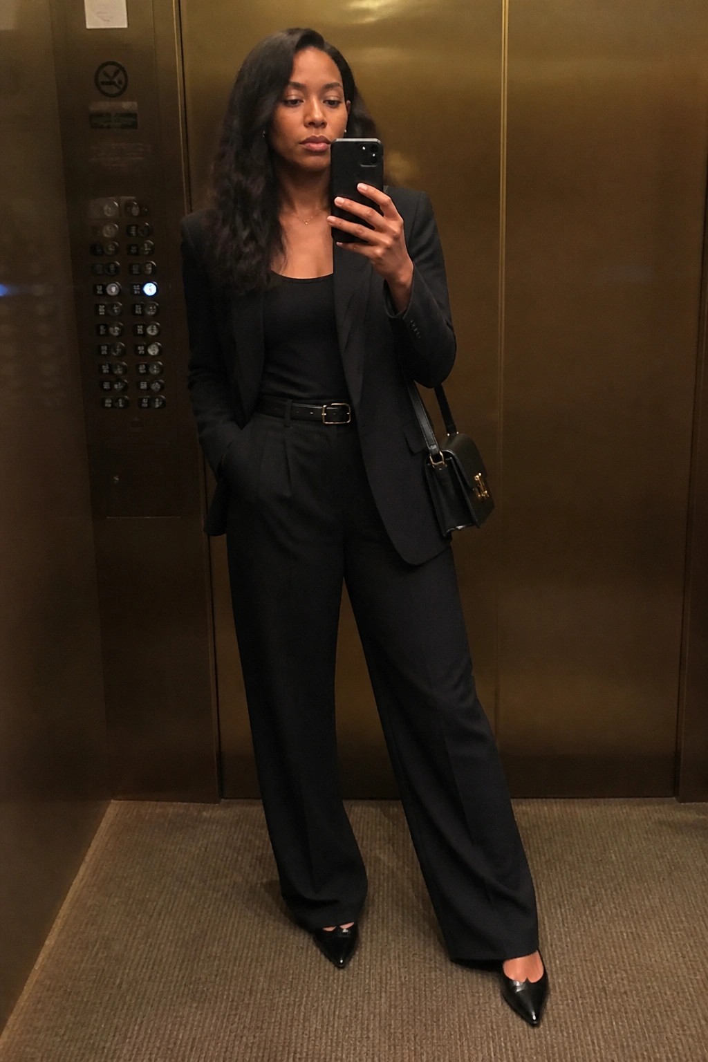 Woman in black pantsuit taking selfie in metallic elevator with handbag