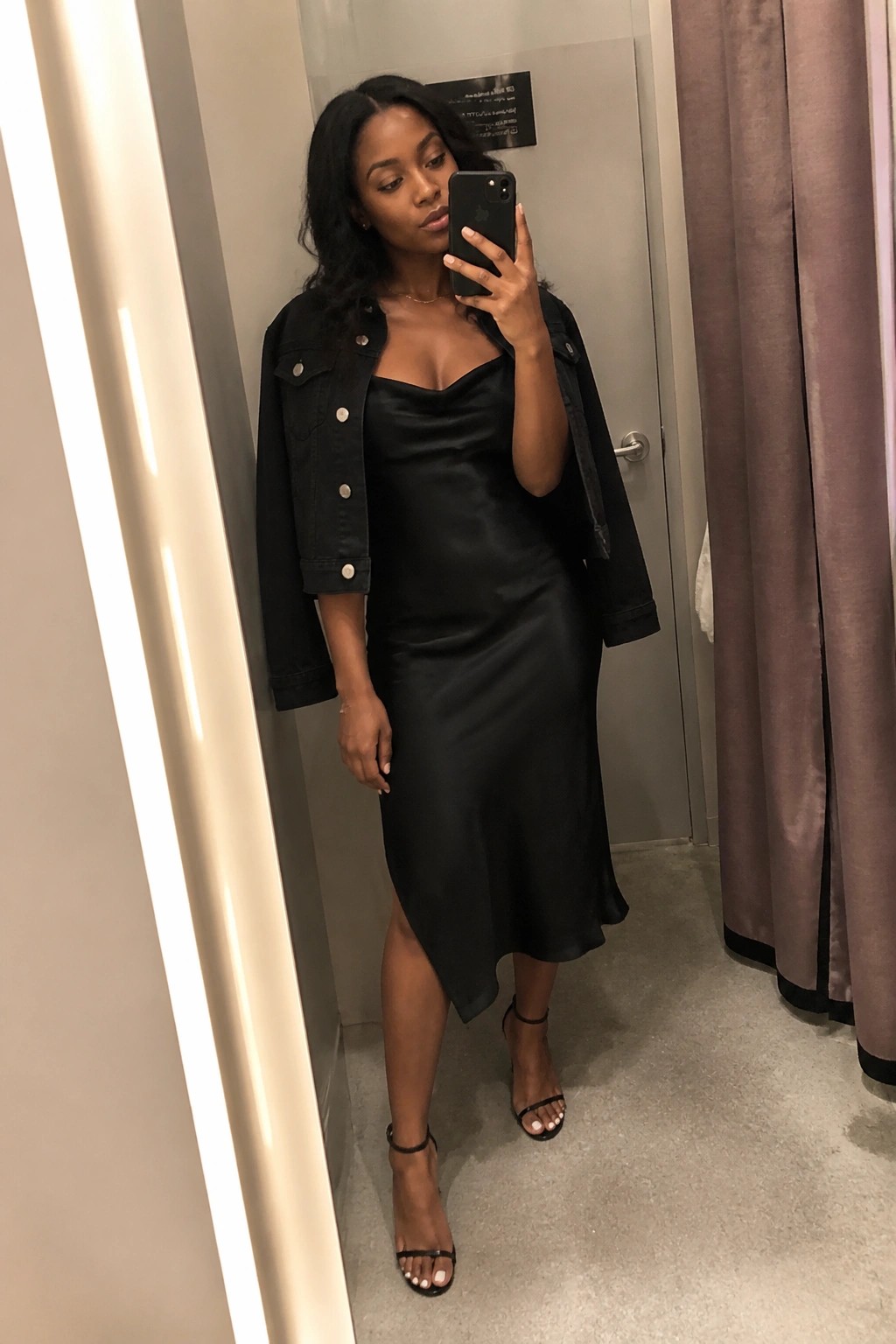Woman wearing a black satin midi slip dress with an asymmetrical hem under a cropped black denim jacket, paired with black strappy heels and a thin gold necklace, taking a mirror selfie in a fitting room.