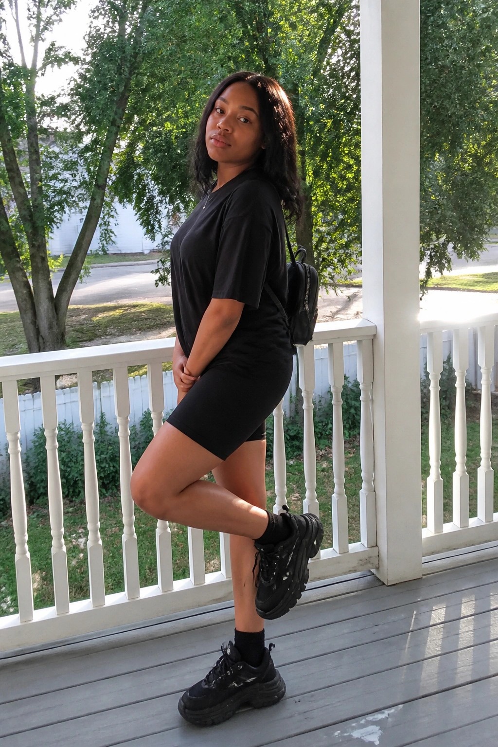Young Black woman in all-black outfit posing on white wooden porch with trees behind