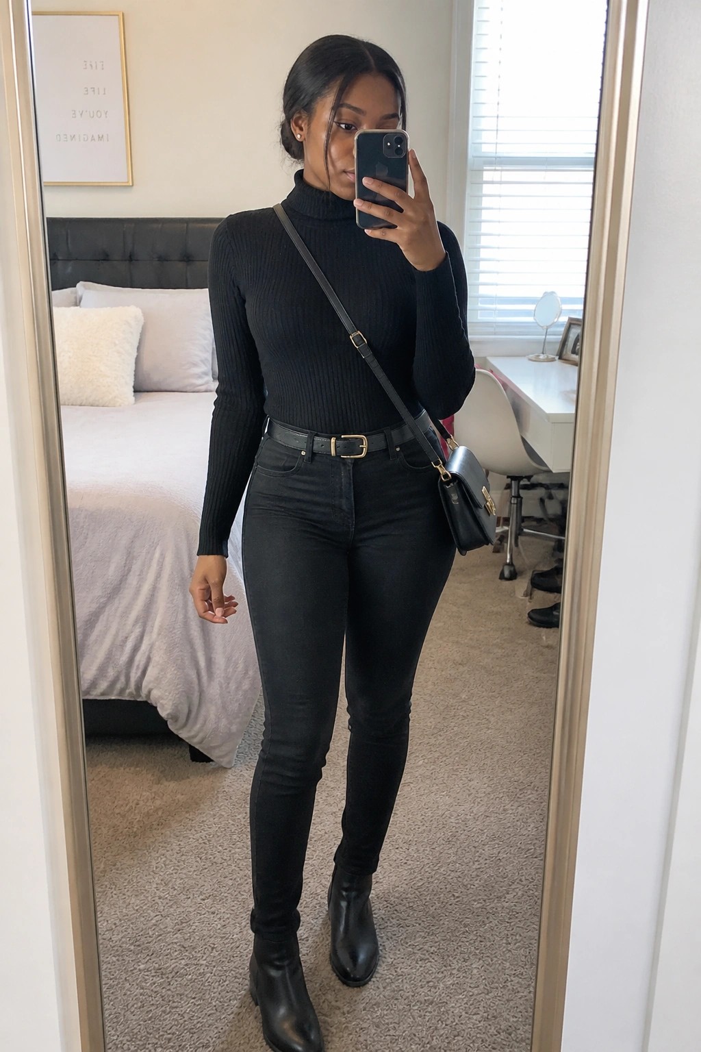 Woman in a mirror selfie wearing an all black ribbed turtleneck sweater tucked into slim black jeans with a gold buckle belt, black ankle boots, and a small black crossbody bag.