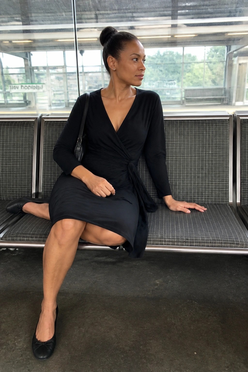 A woman wearing a black long-sleeve wrap dress with a tied waist, black flats, and a black handbag, seated on a bench.
