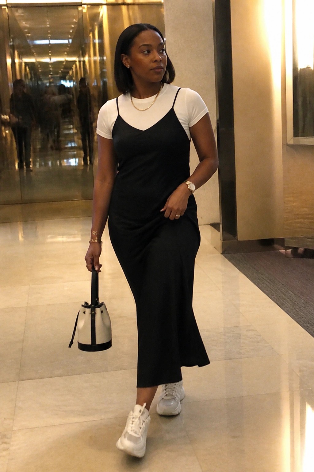 Woman in black slip dress over white tee holding bucket bag in modern hallway