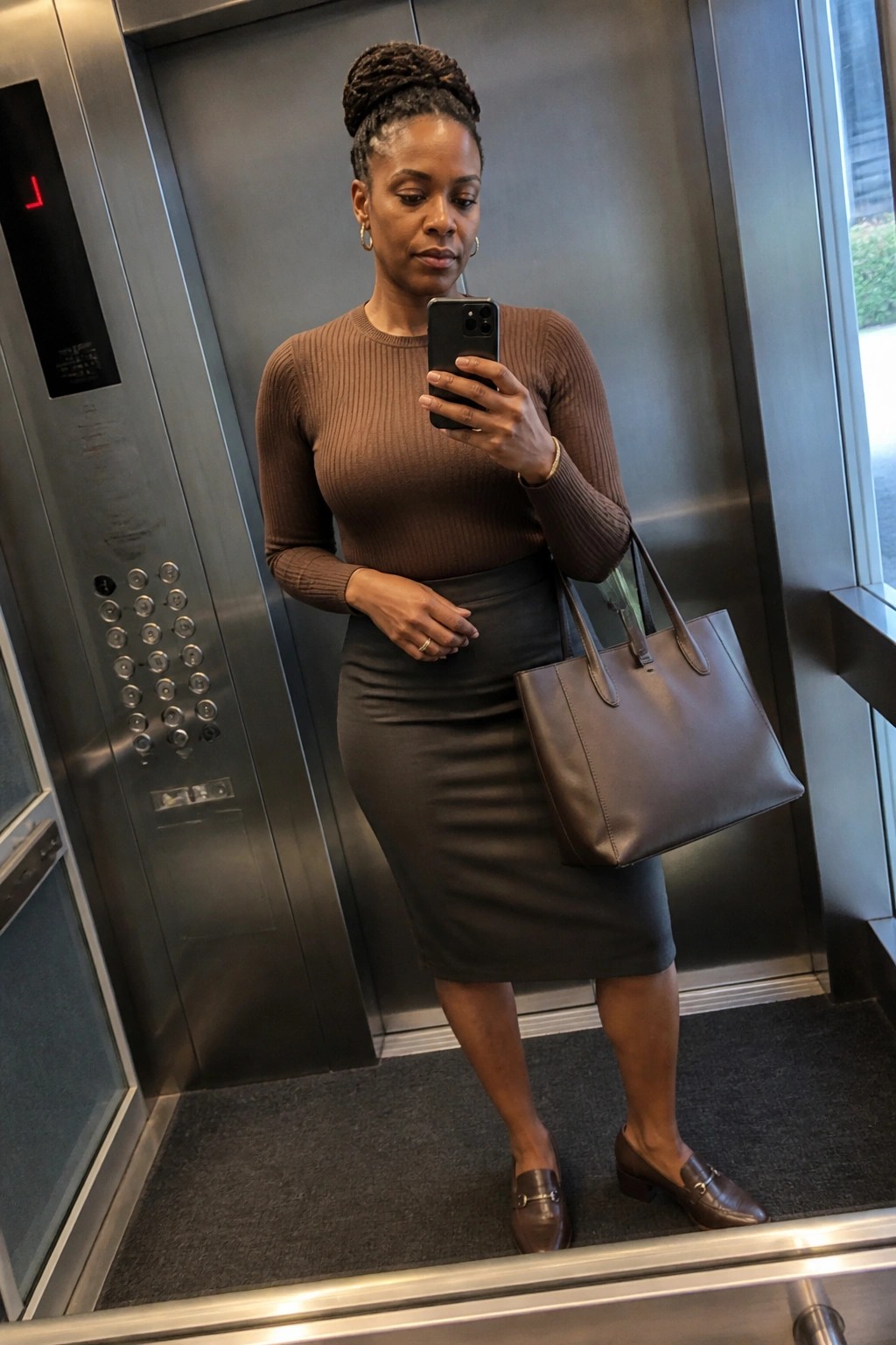 Woman in an elevator taking a mirror selfie while wearing a brown ribbed long sleeve top tucked into a gray pencil skirt with brown loafers and carrying a brown leather tote bag.