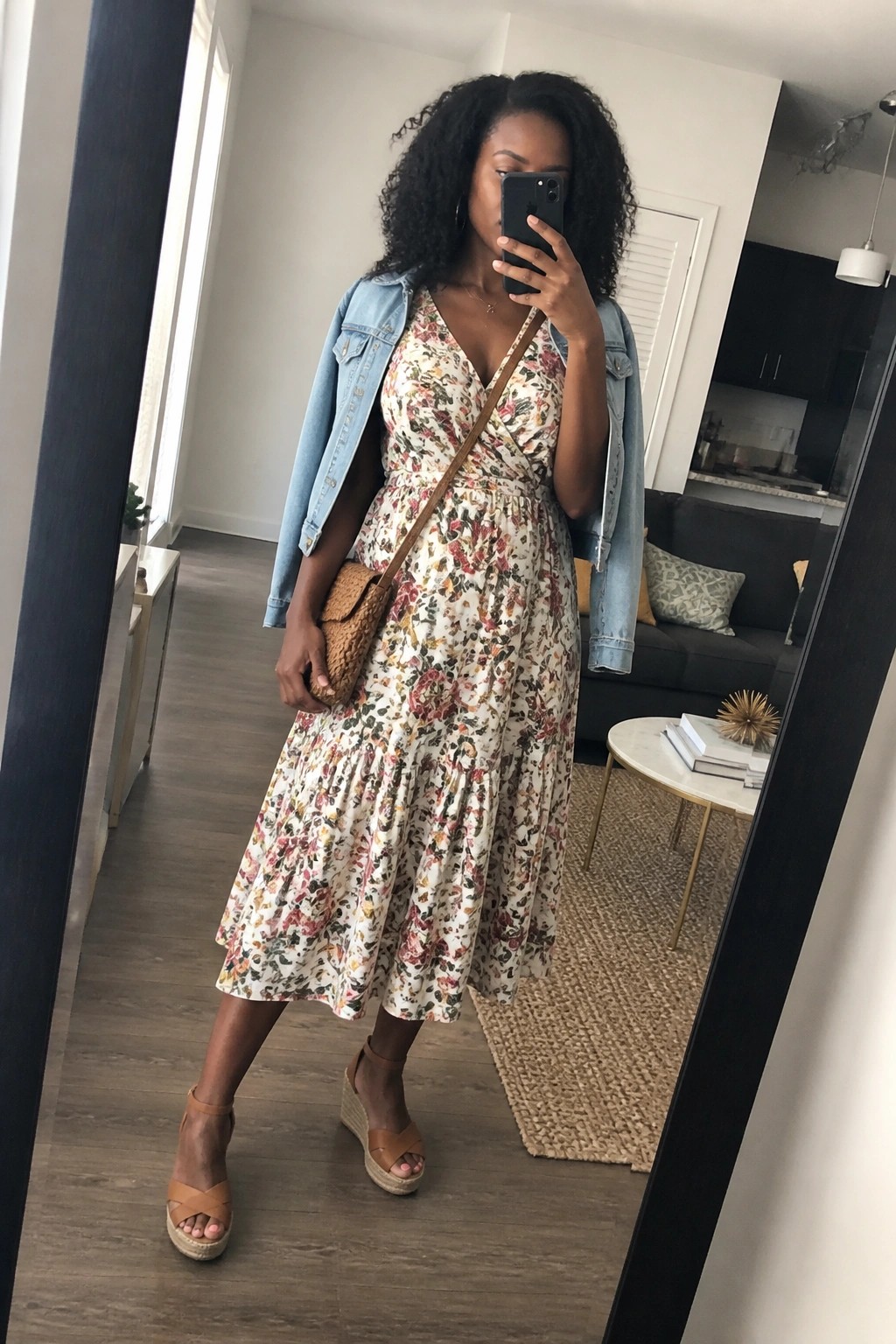 Woman in floral dress and denim jacket taking mirror selfie in apartment