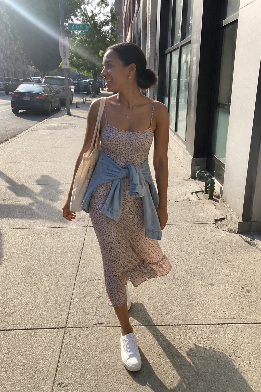 Woman in floral dress with tied denim jacket walks sunny city sidewalk