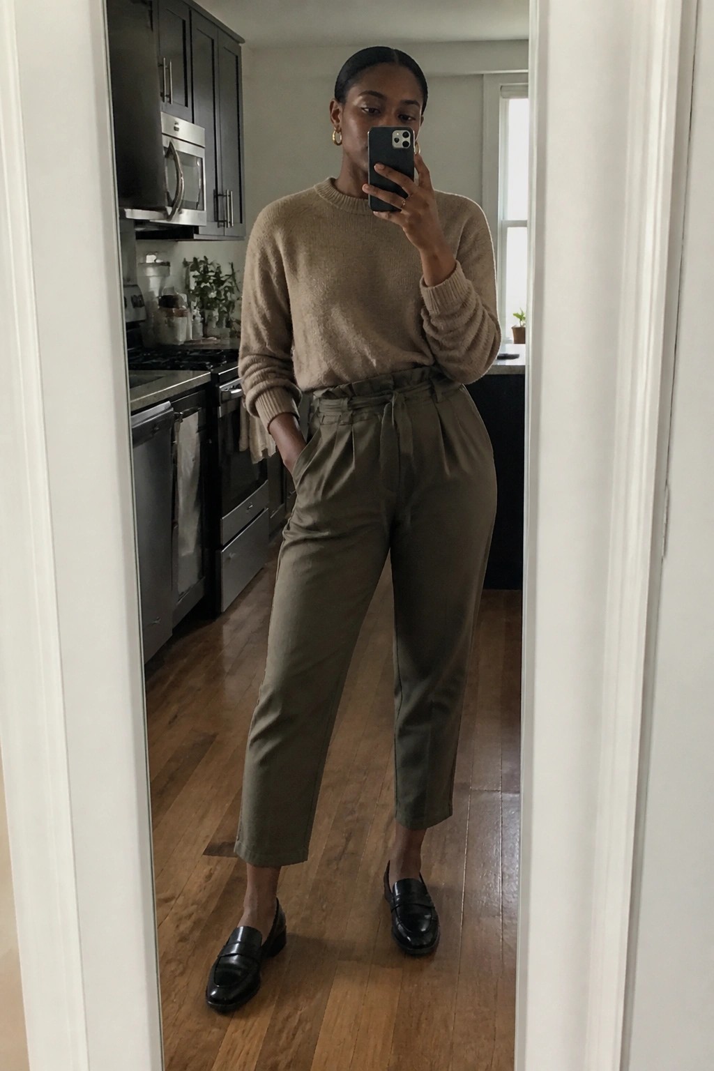 Woman in beige sweater and olive pants taking mirror selfie in kitchen.