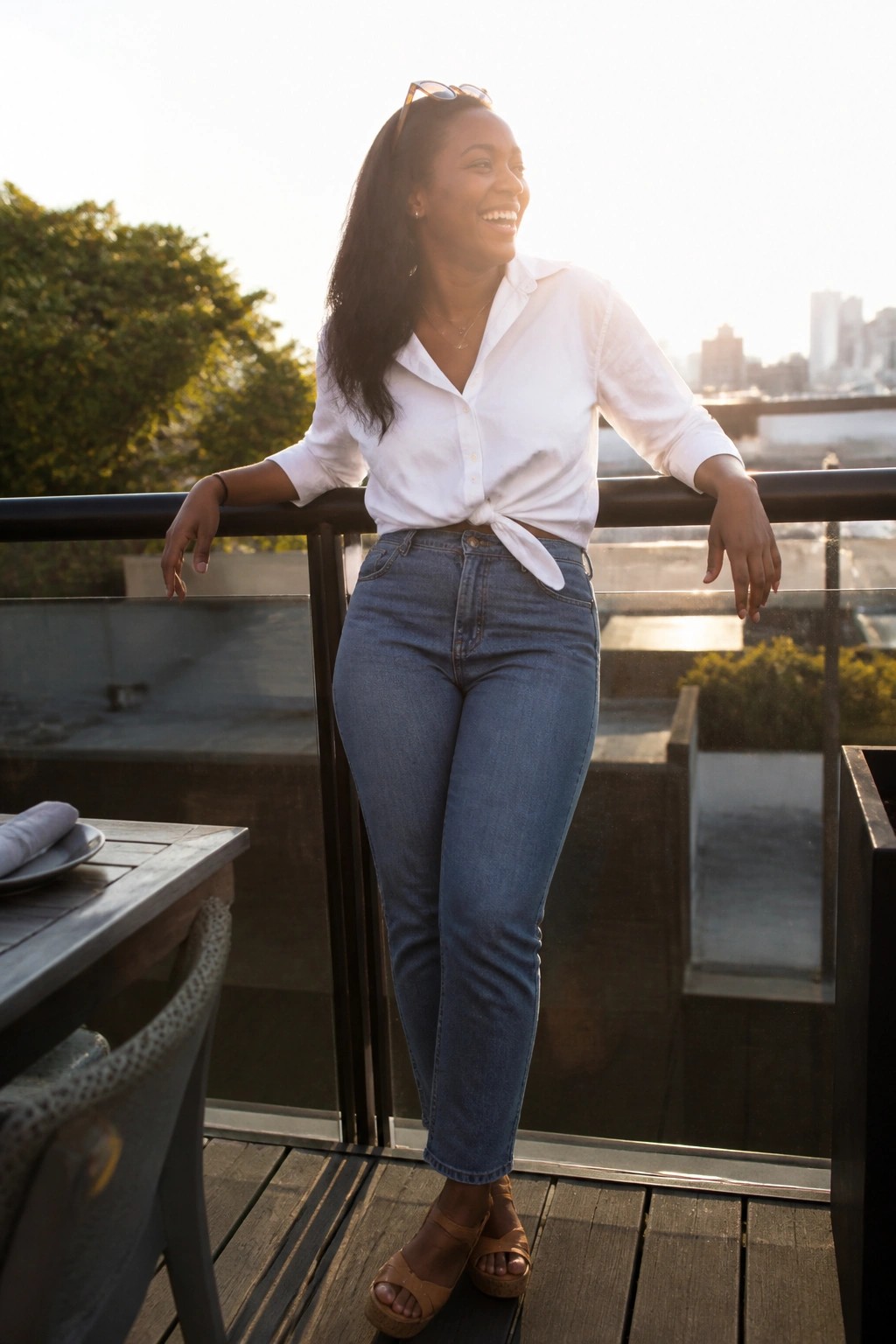 Woman wearing a white button down shirt tied in a knot at the waist over blue slim jeans with brown platform sandals and minimal gold jewelry.