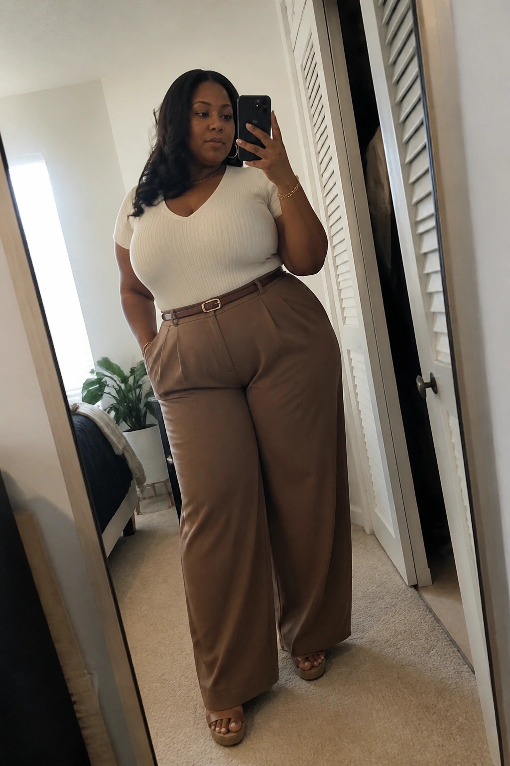Curvy woman in white ribbed top and brown wide-leg pants taking mirror selfie