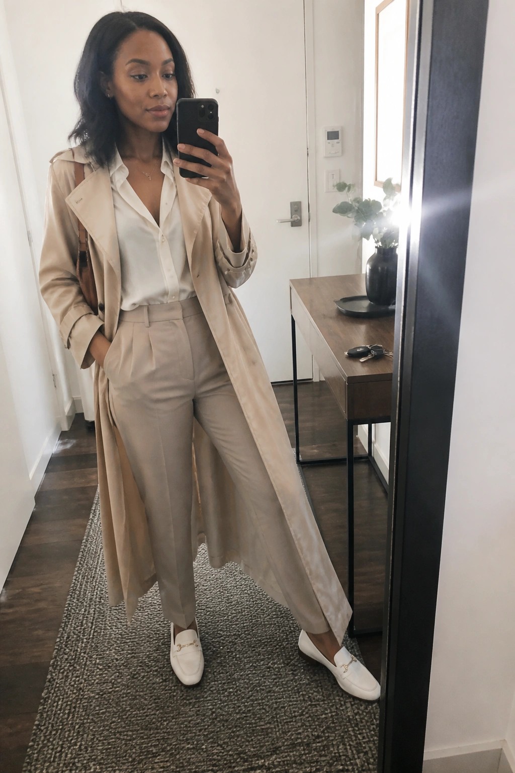 Woman in beige trench coat and trousers taking mirror selfie indoors.