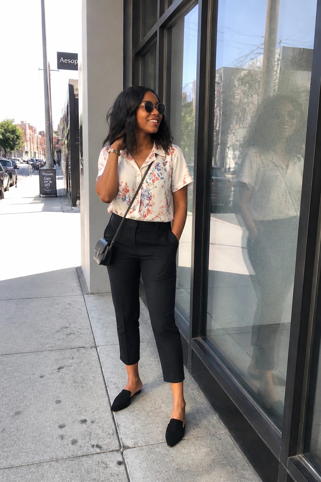 Woman in floral blouse and black pants smiling on city sidewalk by glass window