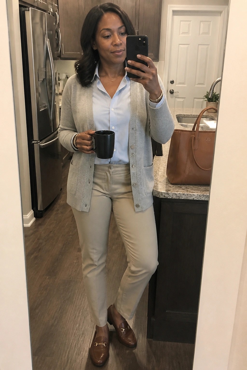 Woman wearing a light gray cardigan over a light blue button down shirt with beige pants and brown loafers holding a black mug in a kitchen setting.