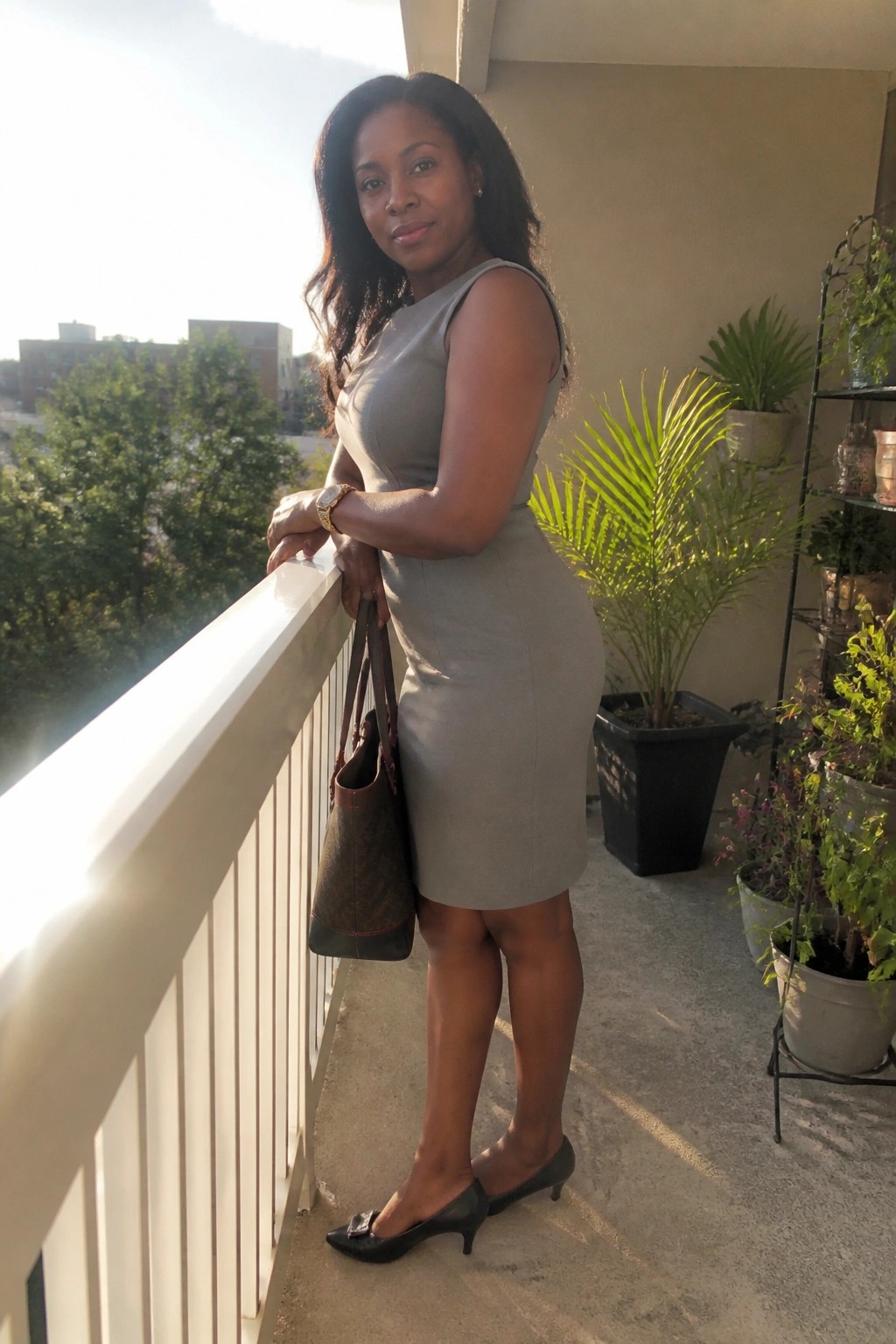 Woman wearing a sleeveless gray sheath dress with black pointed toe heels and holding a patterned handbag on a balcony.